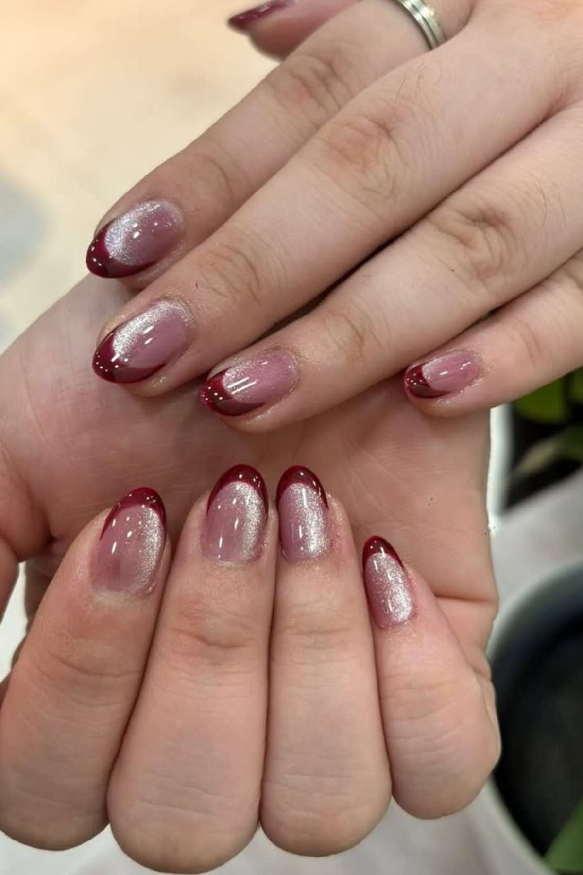 Hands with almond-shaped nails featuring a shimmery pink base and dark red French tips, shown against a neutral background.