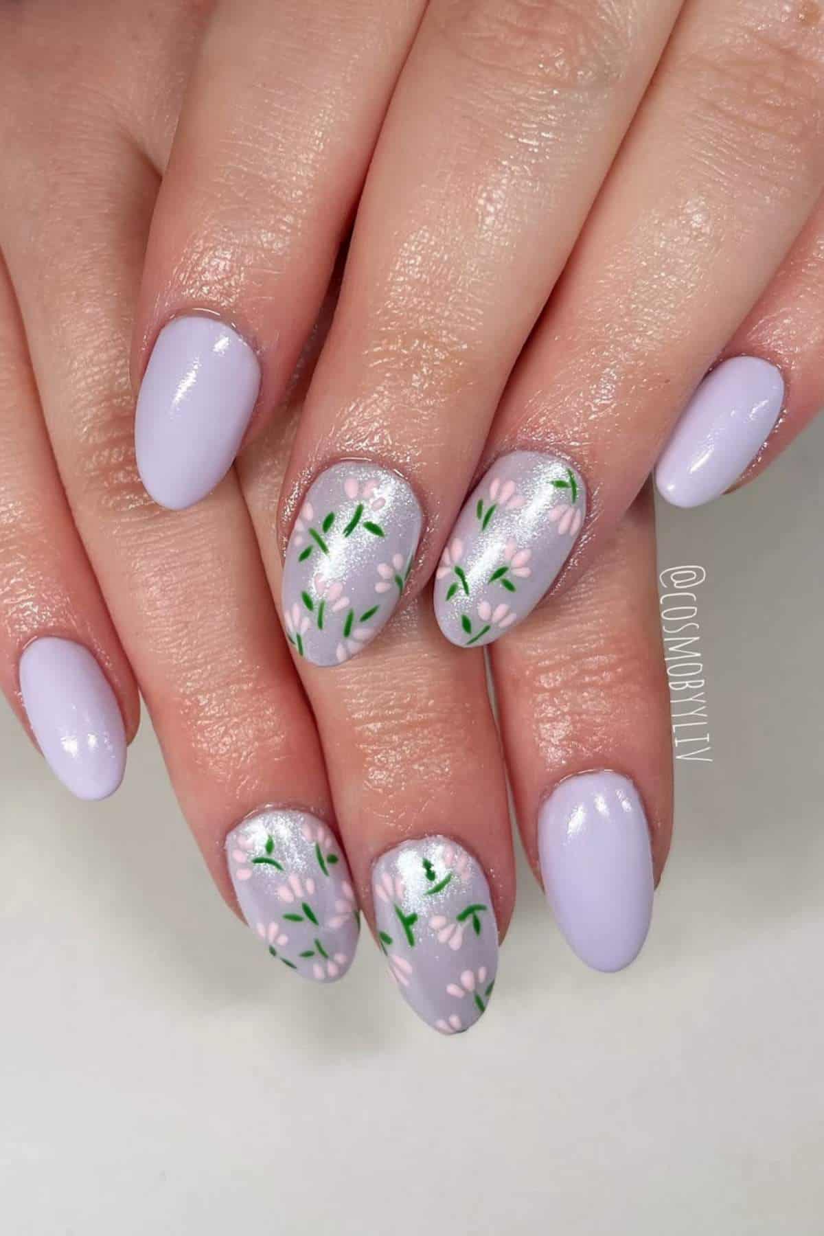 Hands with oval-shaped nails painted light purple; two accent nails on each hand feature a floral design with green leaves and white flowers.