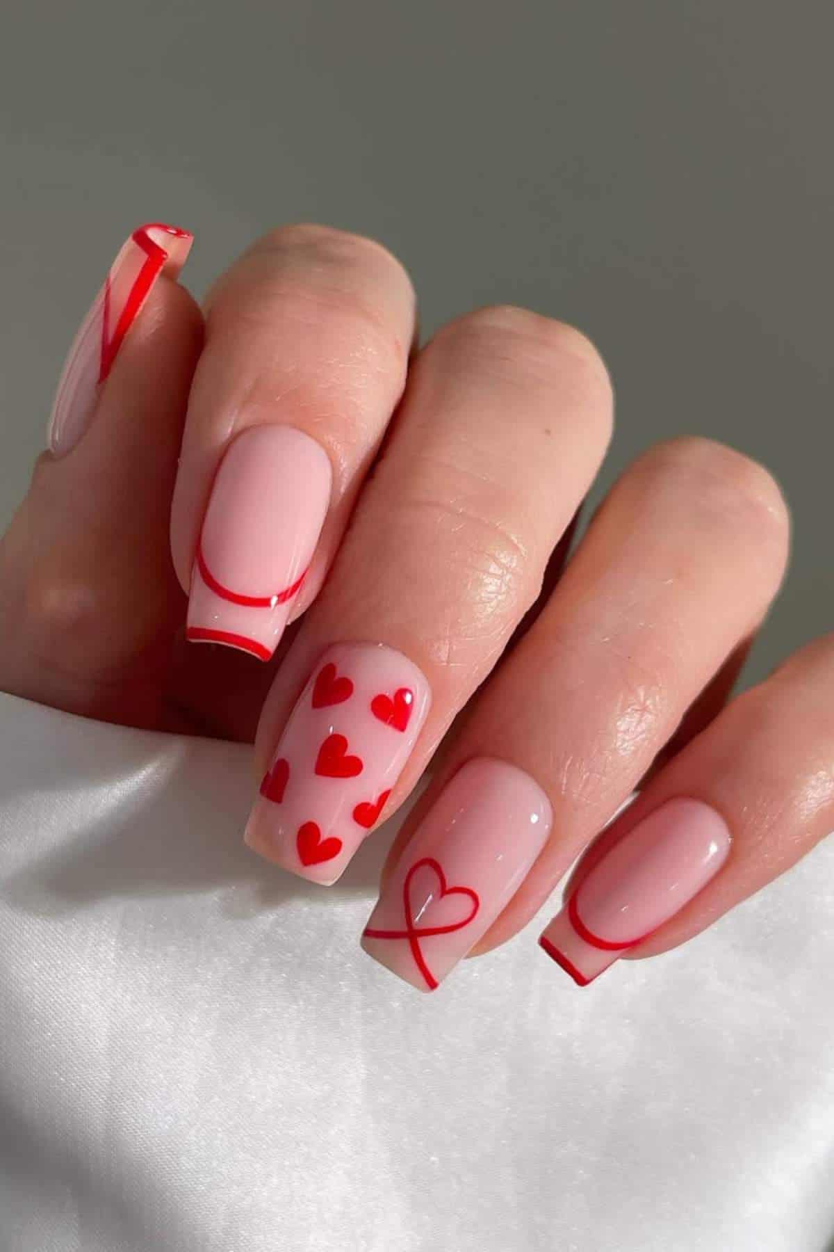 A hand with light pink nails featuring red tips, red heart designs, and a single outlined heart accent nail.