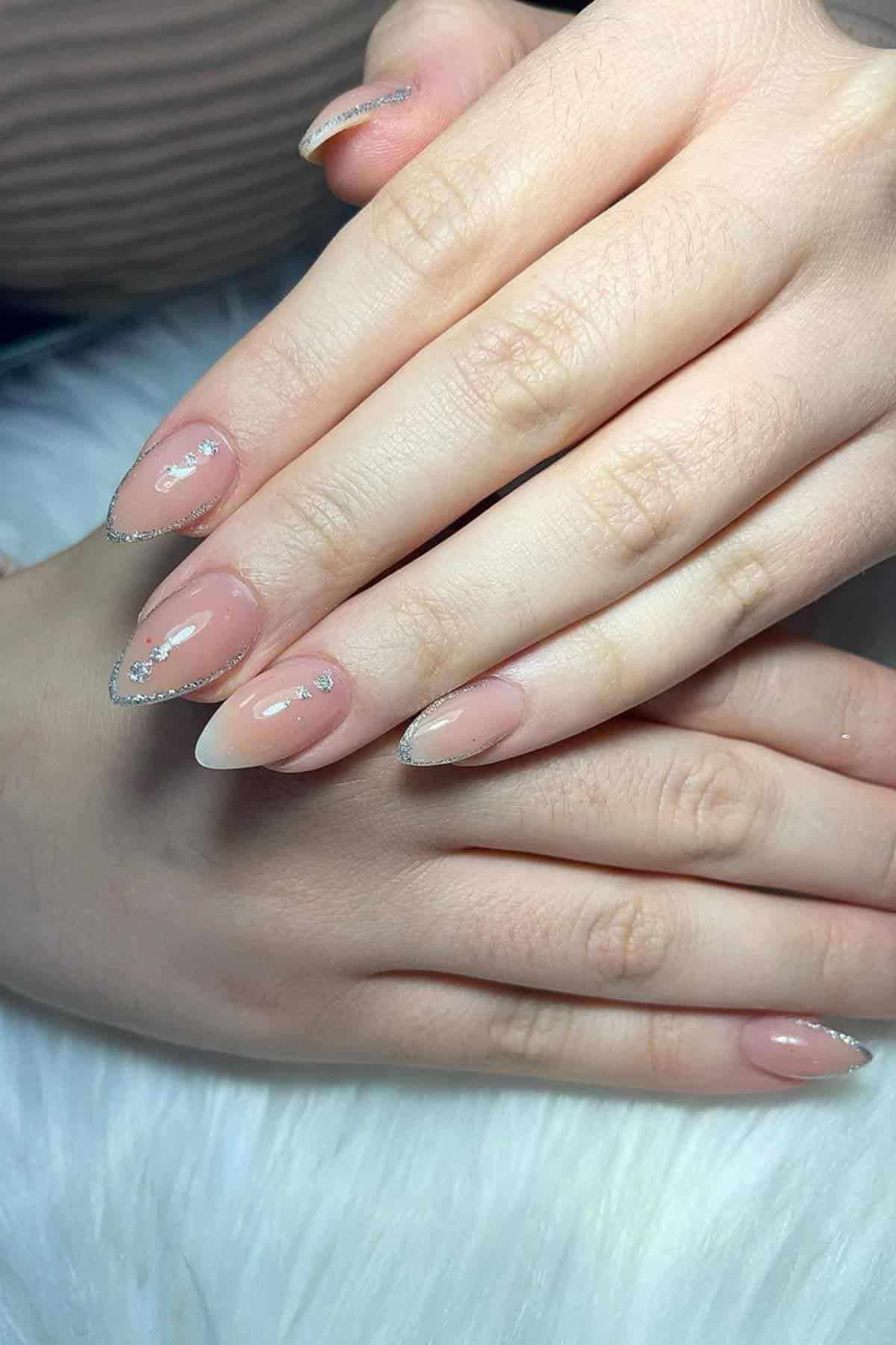 Hands with almond-shaped nails featuring a nude pink base, silver glitter tips, and small decorative rhinestones, resting on a white surface.