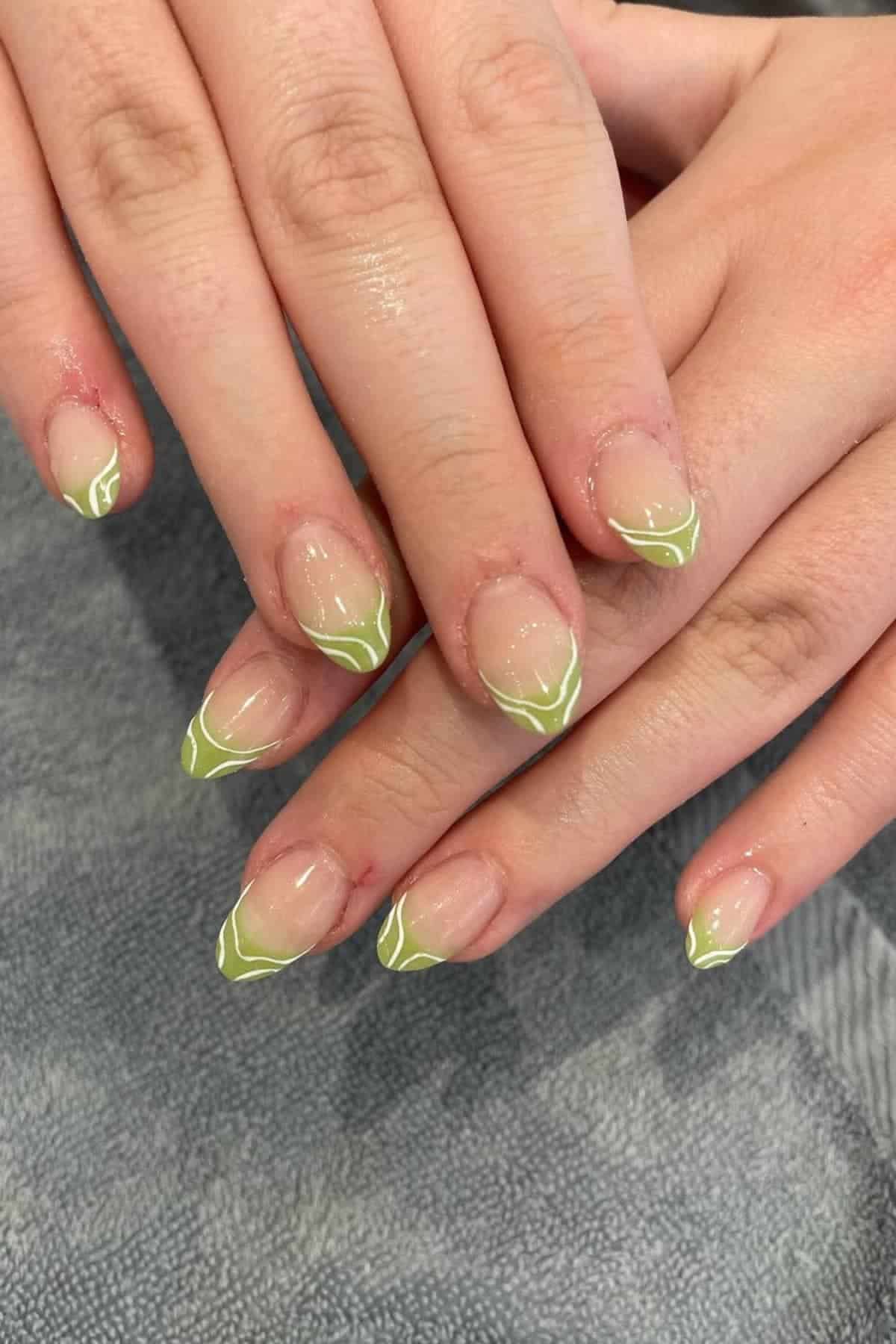 Hands with almond-shaped nails featuring a light green French tip design outlined with a thin white line, resting on a gray textured surface.