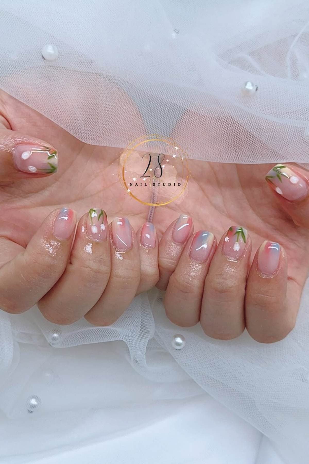 Close-up of hands with short, oval-shaped nails featuring a clear base and green-tipped French manicure with small white flower accents.