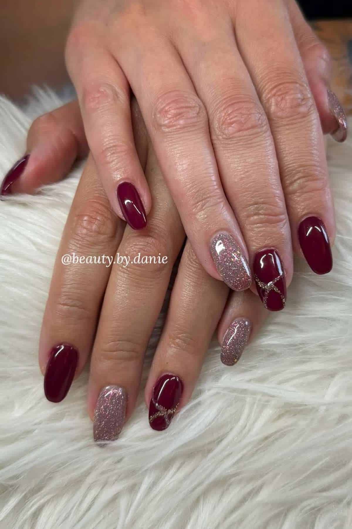 Hands with manicured nails featuring burgundy polish and glittery accent nails, some with gold decorative lines, resting on a white textured surface.