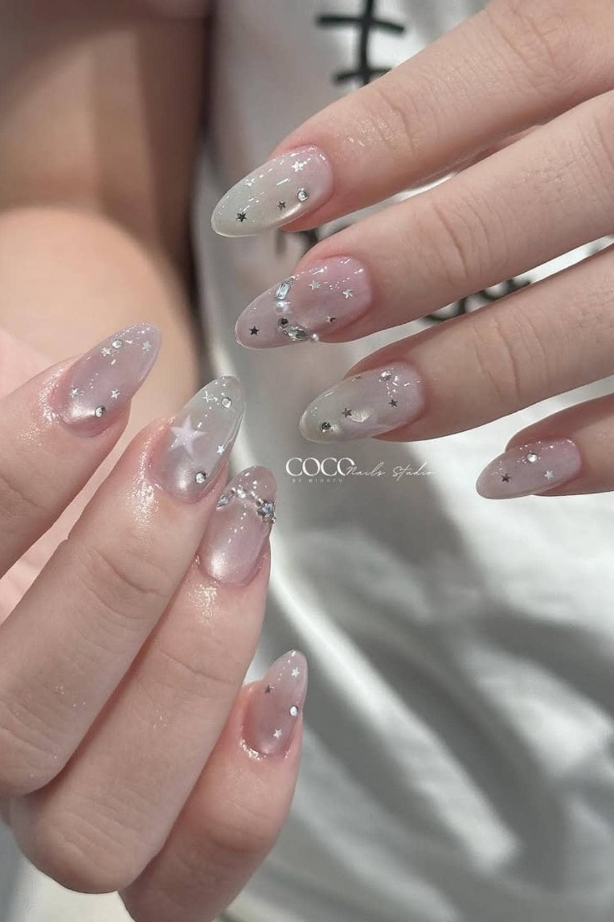 A close-up of hands with long, almond-shaped nails featuring a translucent base and decorated with silver stars and small rhinestones.