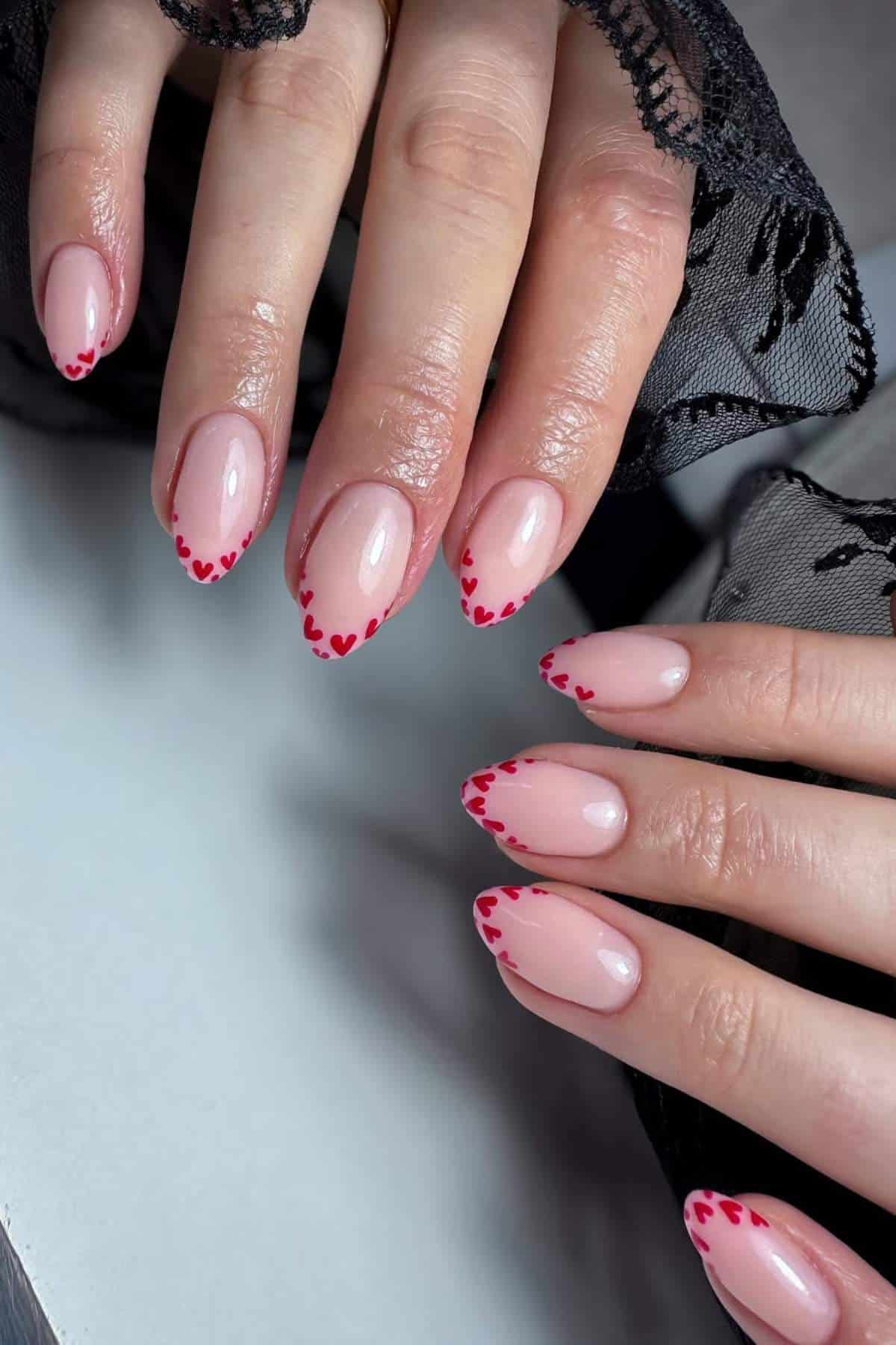 Hands with almond-shaped nails featuring a pale pink base and small red heart designs along the tips, partially covered by black lace fabric.