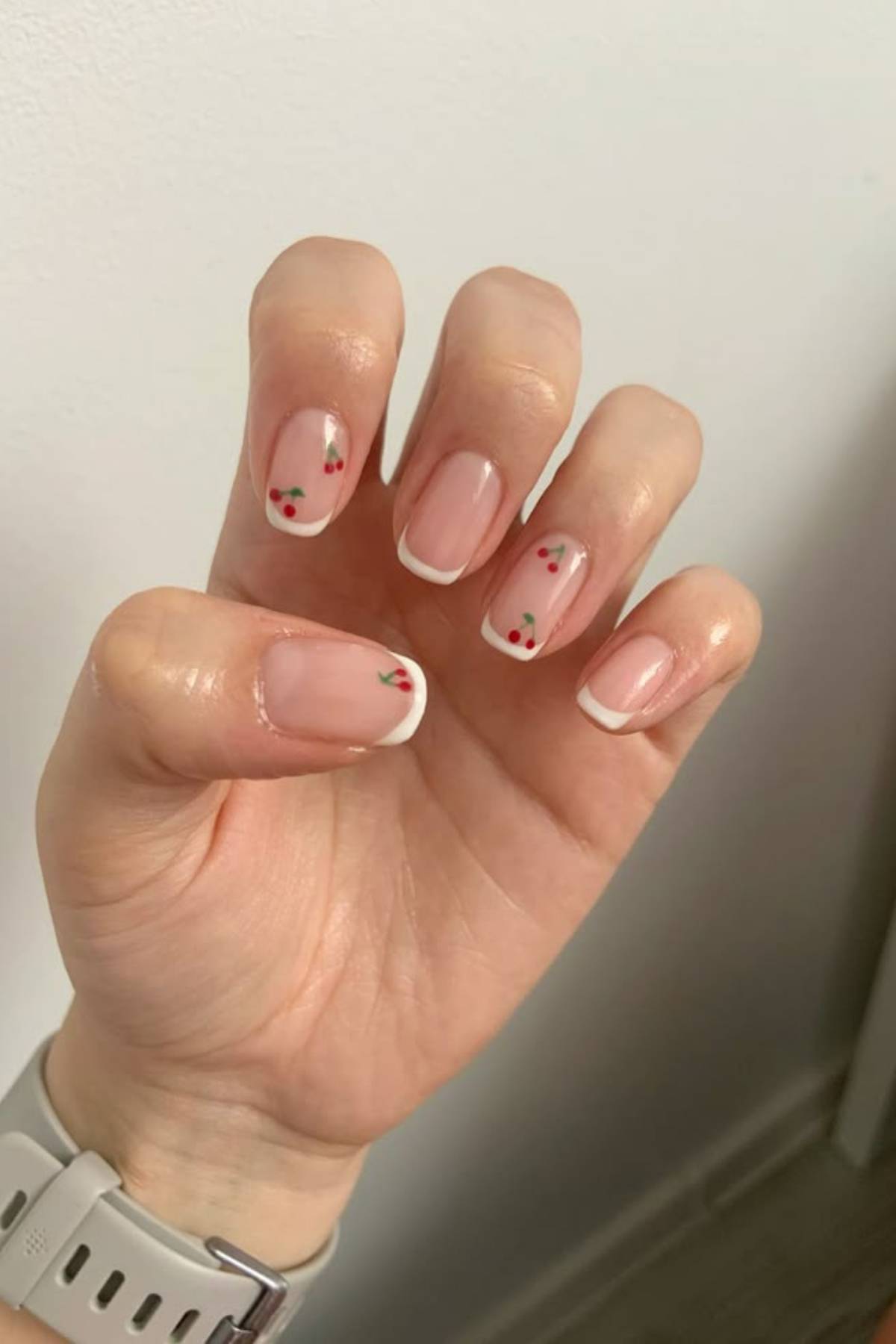 A hand with a white smart watch shows nails with a French manicure and small red cherry designs on some nails.