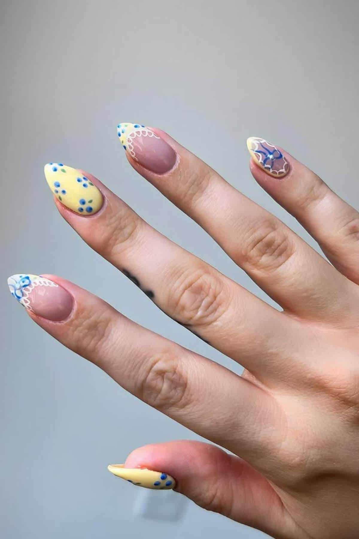 A hand with almond-shaped nails features yellow, blue, and white abstract nail art designs on a neutral background.