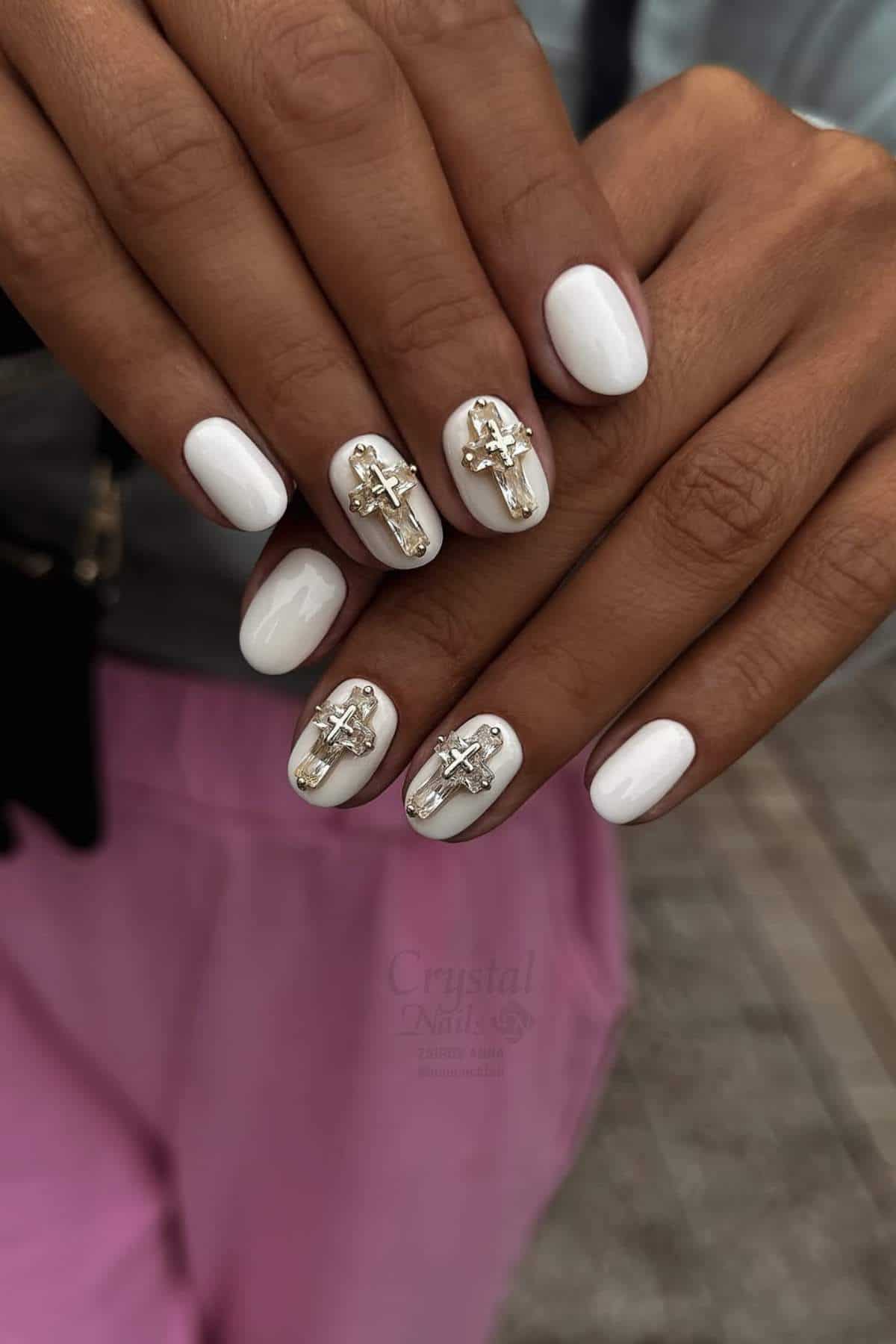 A hand with white oval-shaped nails featuring gold accents and metallic cross-shaped decorations on each nail.