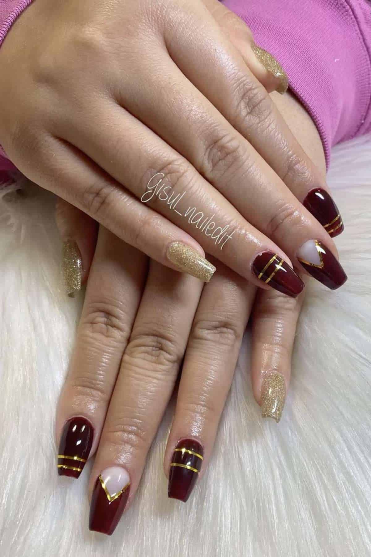 Hands with manicured nails featuring burgundy polish with gold lines, nude accents, and gold glitter polish on ring fingers, resting on a white surface.