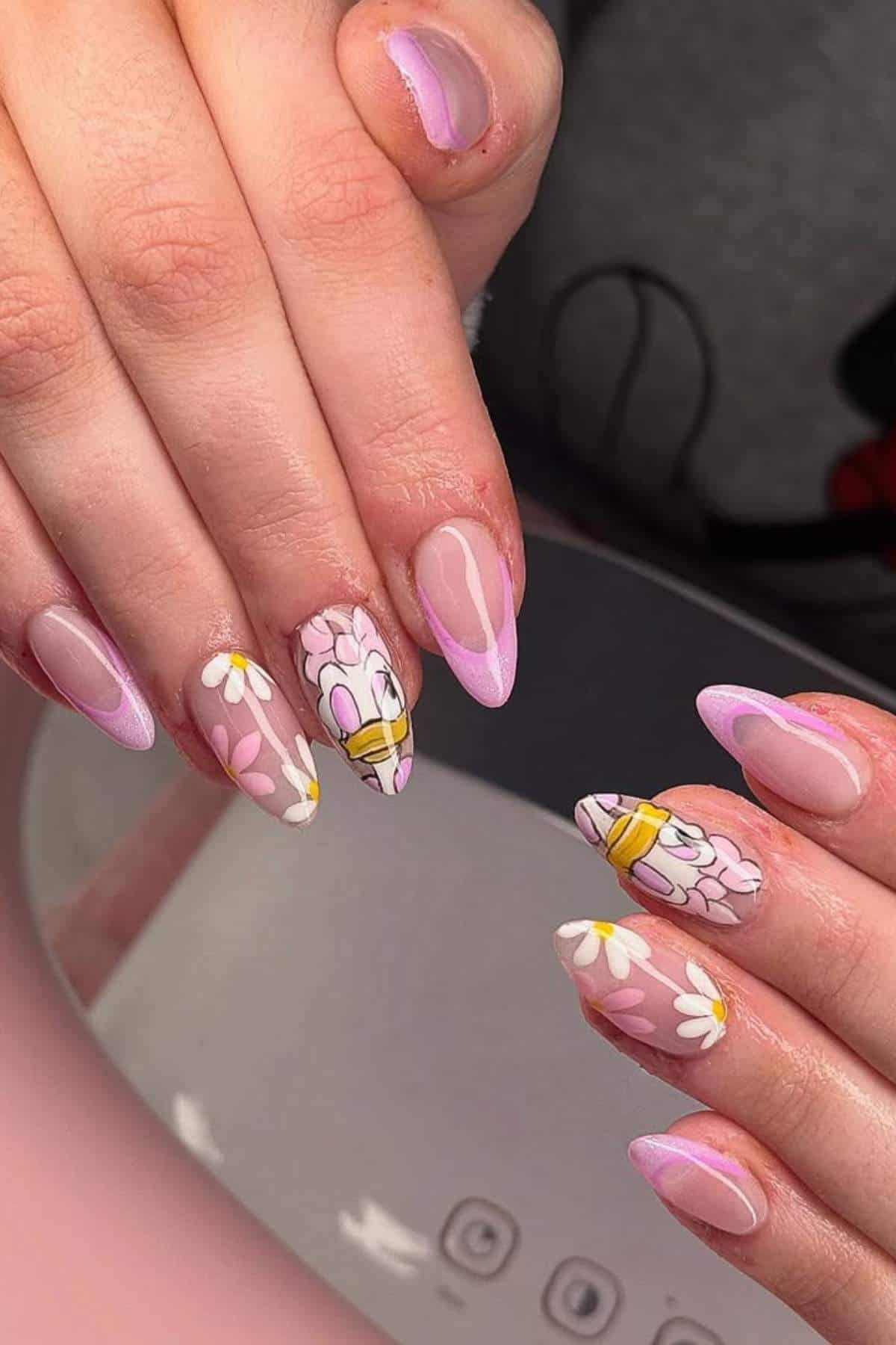 Hands with long, almond-shaped nails featuring pink French tips, daisy flower designs, and cartoon character art on two accent nails.