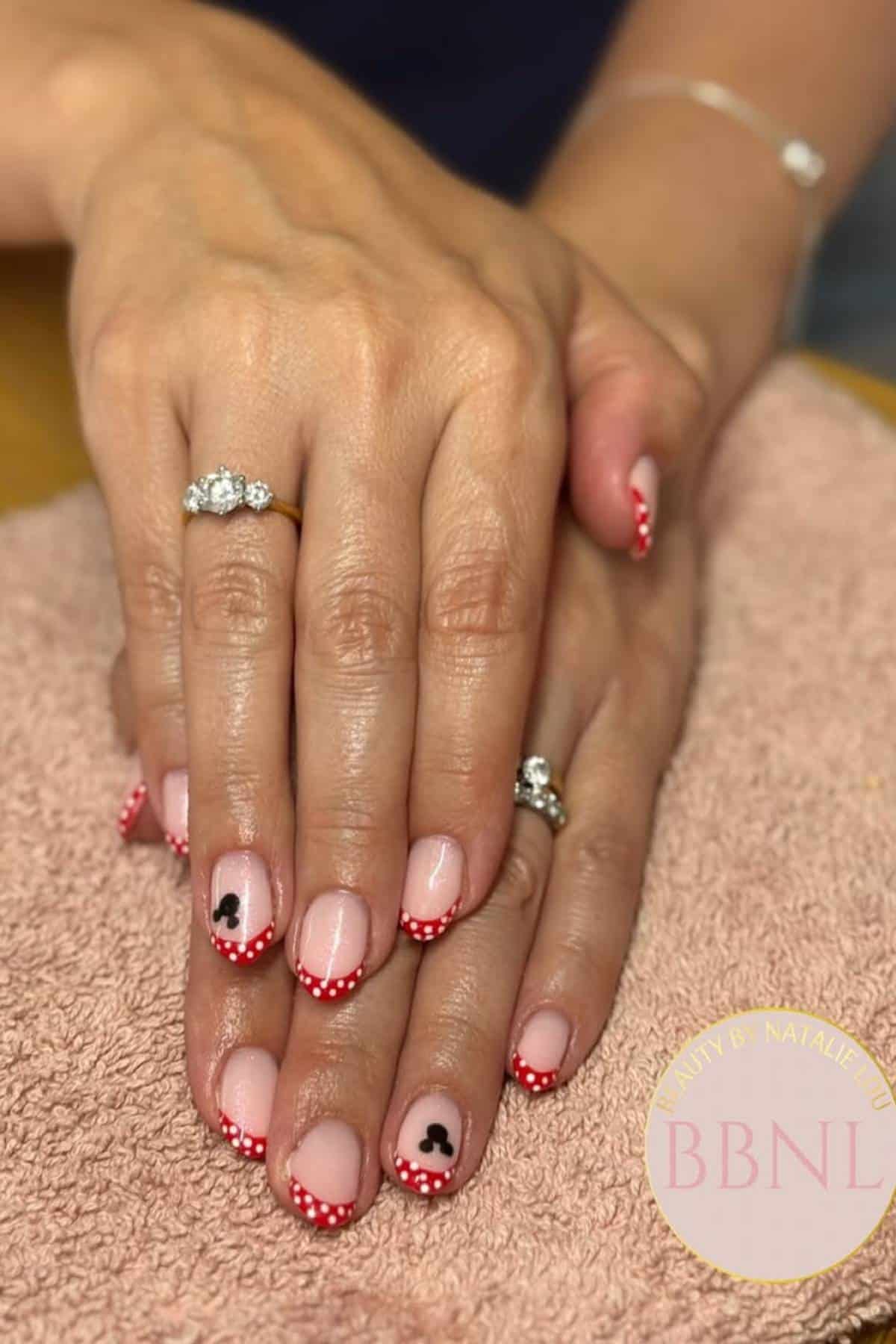 Hands with a pink and red polka dot French manicure, featuring small black Mickey Mouse designs on the ring fingernails, resting on a light brown towel.