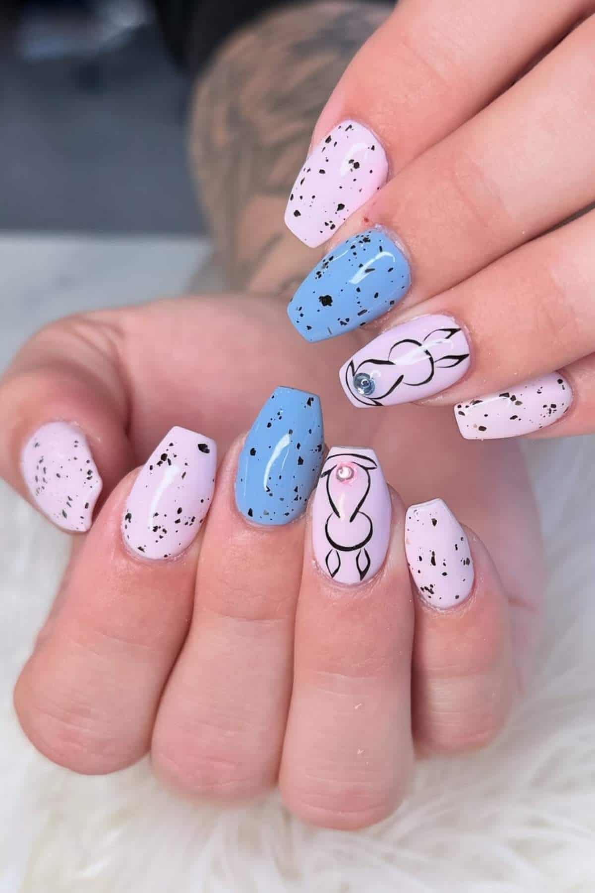 A hand with square-shaped nails painted in pastel pink and blue, featuring black speckles and abstract black line art designs on two nails.