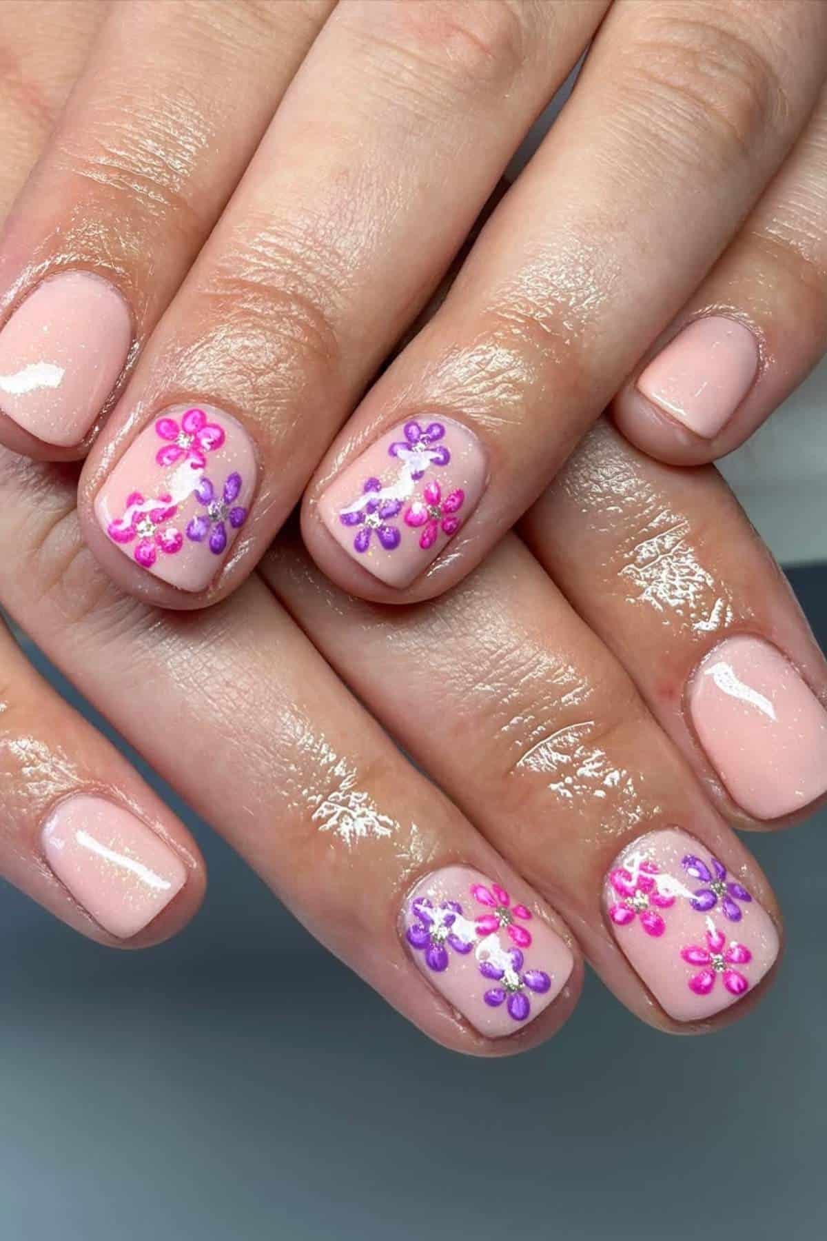 Close-up of hands with short, glossy, light pink nails featuring pink and purple flower designs on the middle and ring fingernails.