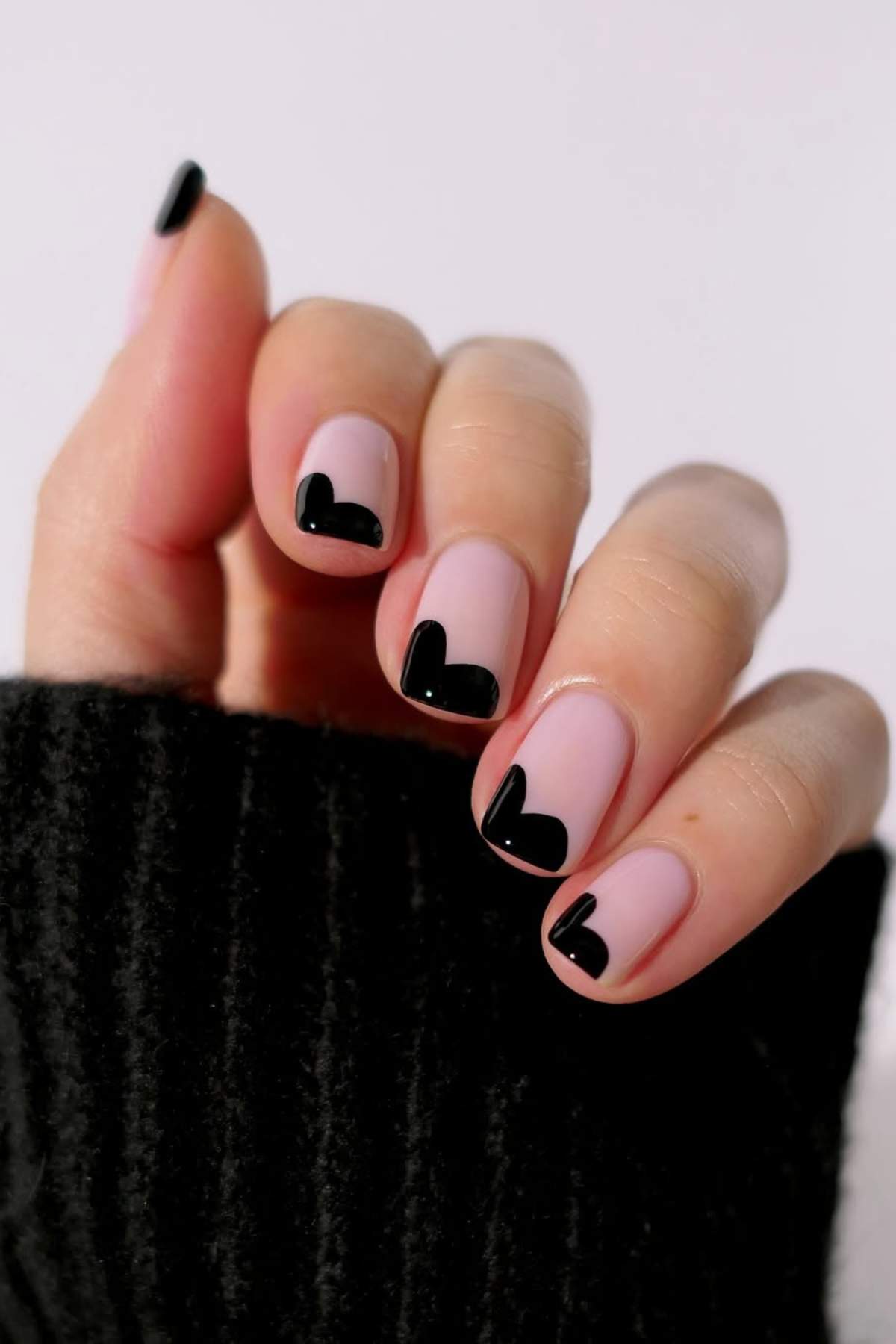 A hand with short nails painted light pink, each featuring a black heart-shaped tip design. The person is wearing a black ribbed sleeve.