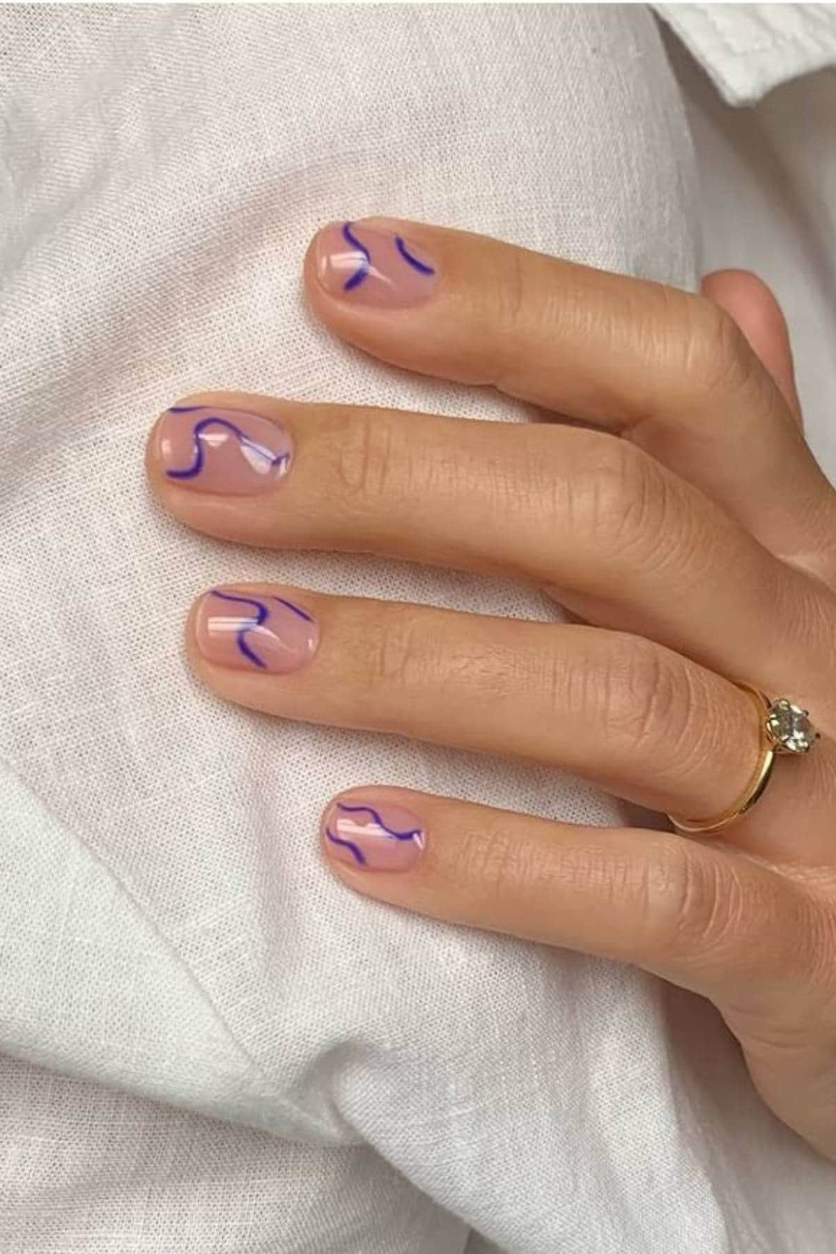 A hand with short, natural nails featuring abstract purple and lavender line designs. The person is wearing a gold ring with a single gemstone.