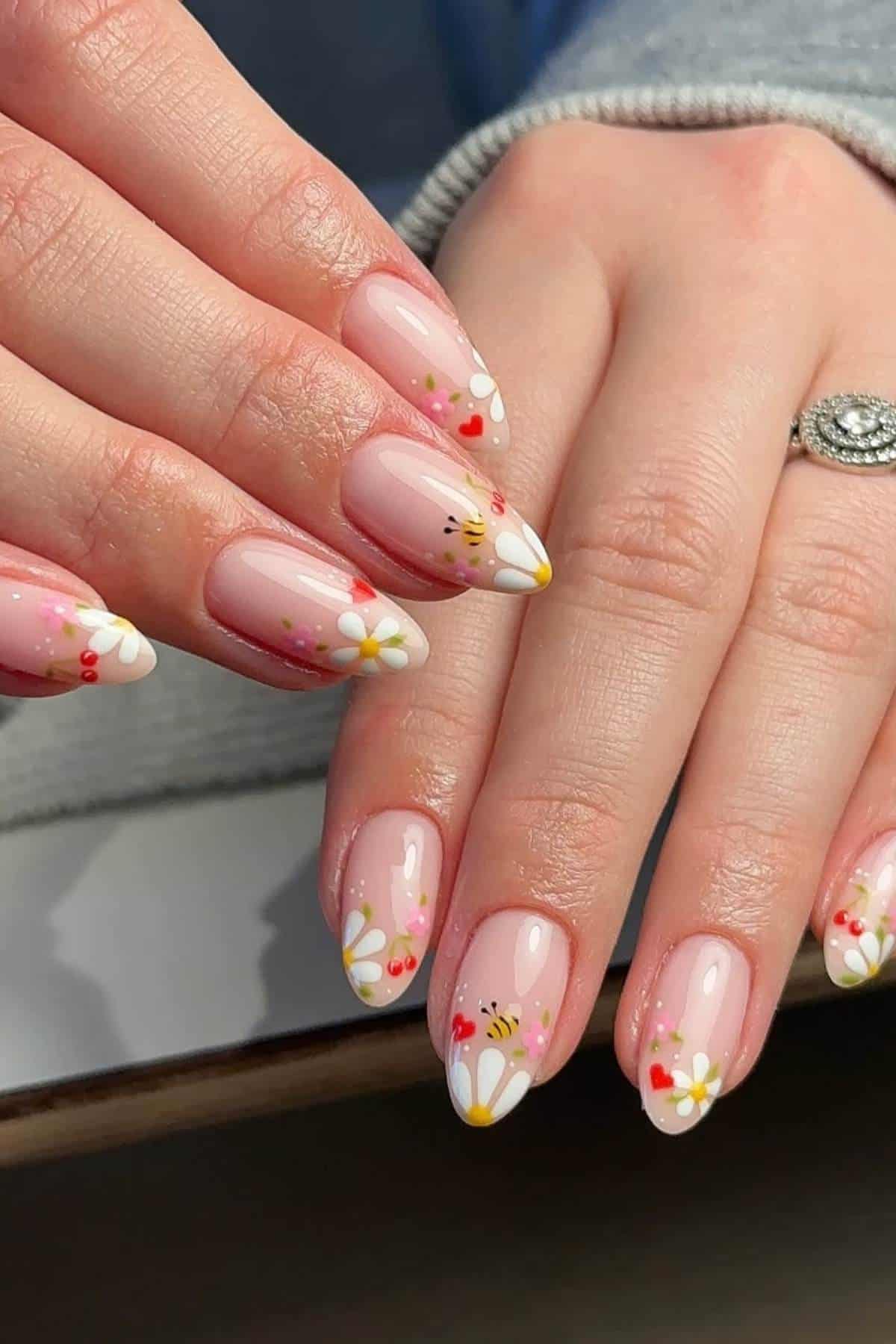 Hands with almond-shaped nails painted light pink, featuring daisy flower designs and small red accents. A silver ring with a large stone is worn on one finger.