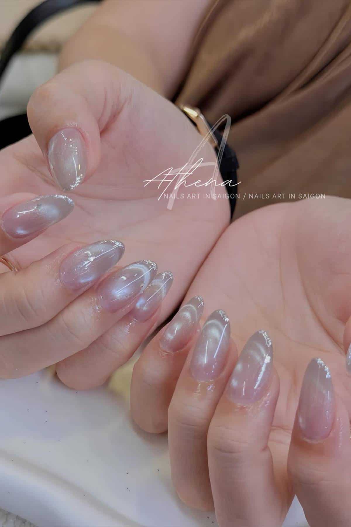 Hands with oval-shaped nails featuring a translucent, shimmery design and silver glitter accents on the tips.