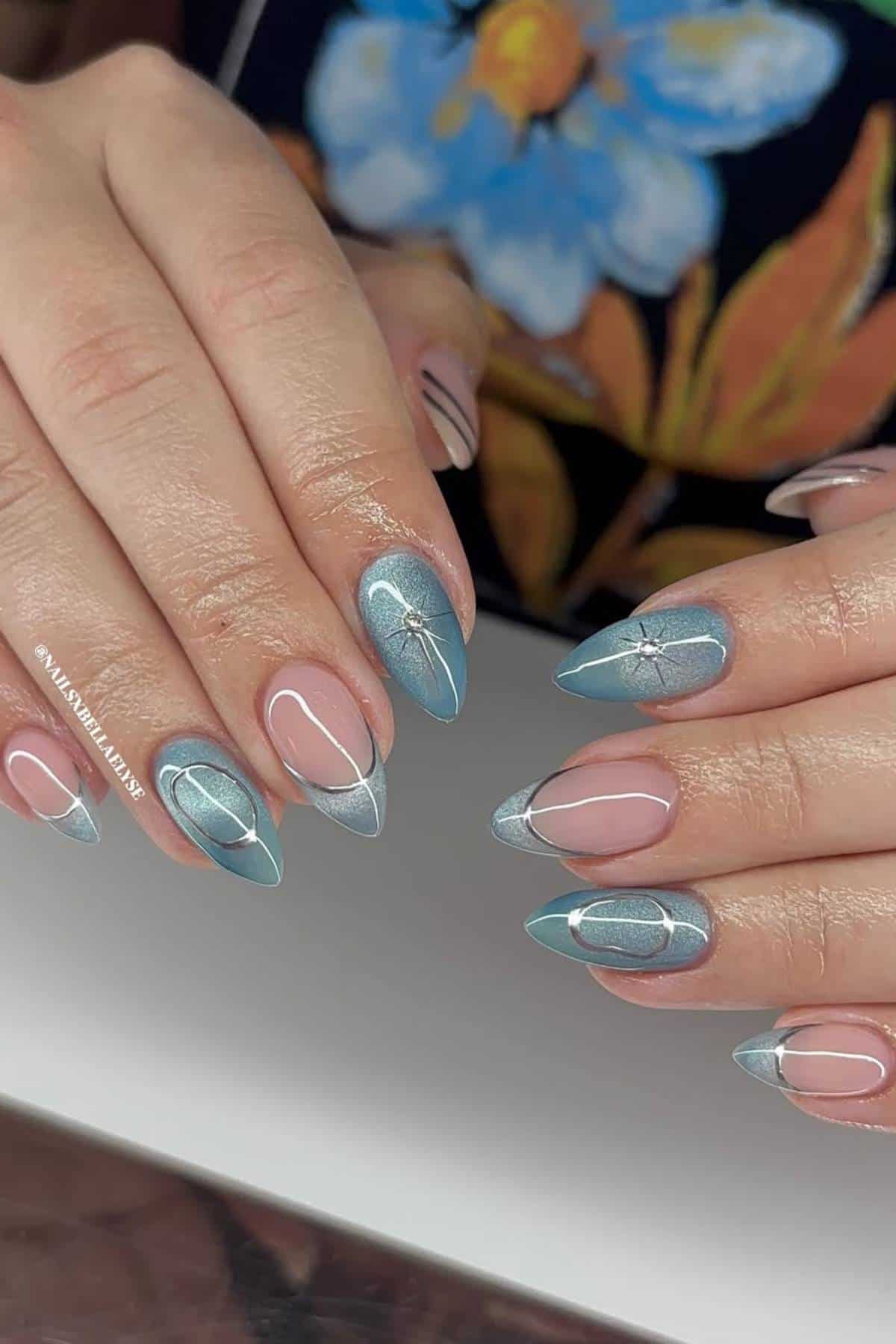 A close-up of hands with almond-shaped nails featuring blue and silver abstract nail art designs, some with delicate line work and small gem accents.