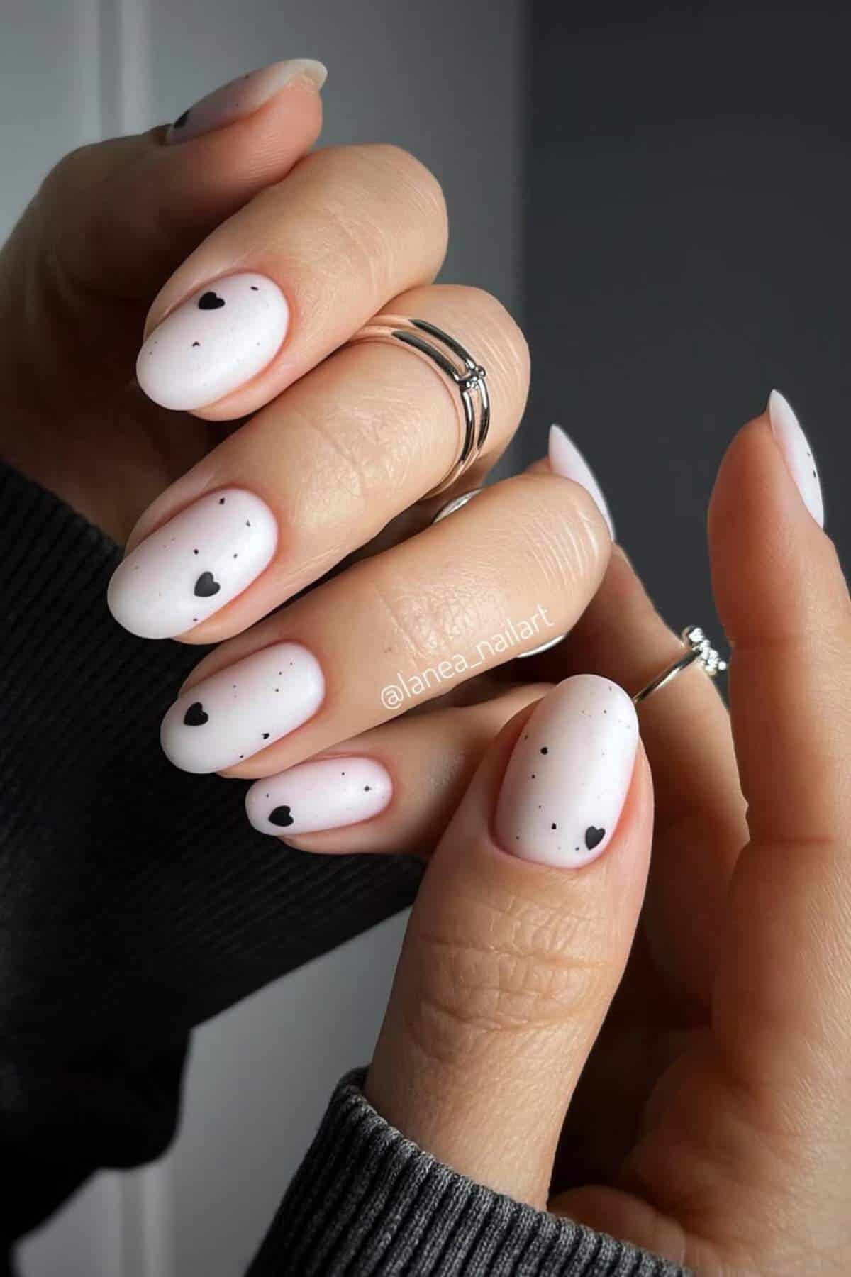 Hands with white matte nail polish, small black hearts and dots as nail art; person is wearing silver rings and a dark long sleeve.