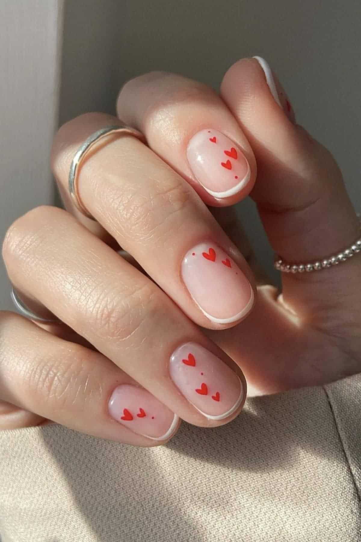 A hand with natural, short nails features a minimalist design of small red hearts on each nail. The person is wearing silver rings on their fingers.