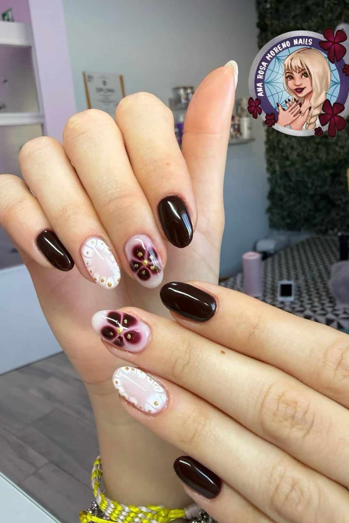 A hand with manicured nails featuring dark maroon polish and accent nails with pink, white, and floral designs, shown in a nail salon setting.