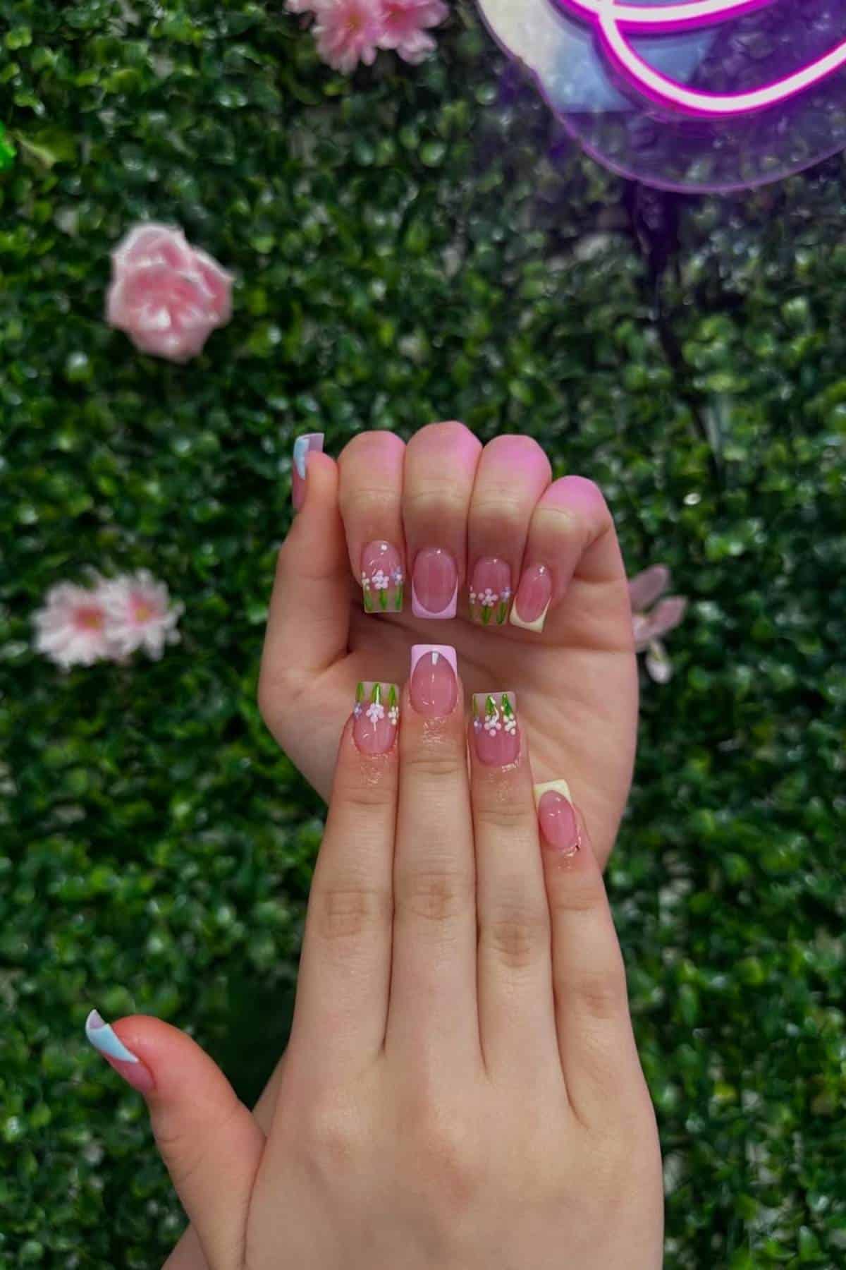 Hands displaying manicured nails with pink polish and white flower designs, set against a green leafy background with pink flowers.