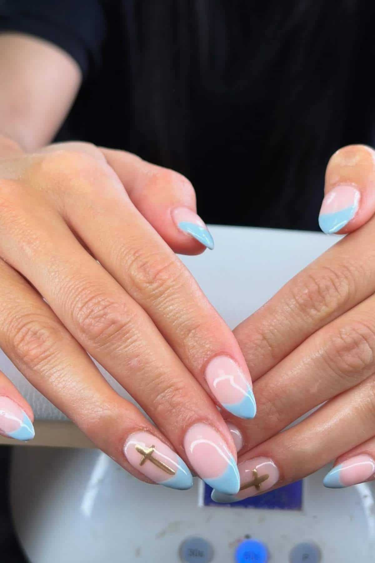 Hands with almond-shaped nails featuring a light blue French tip design and gold cross decals on the ring fingers.