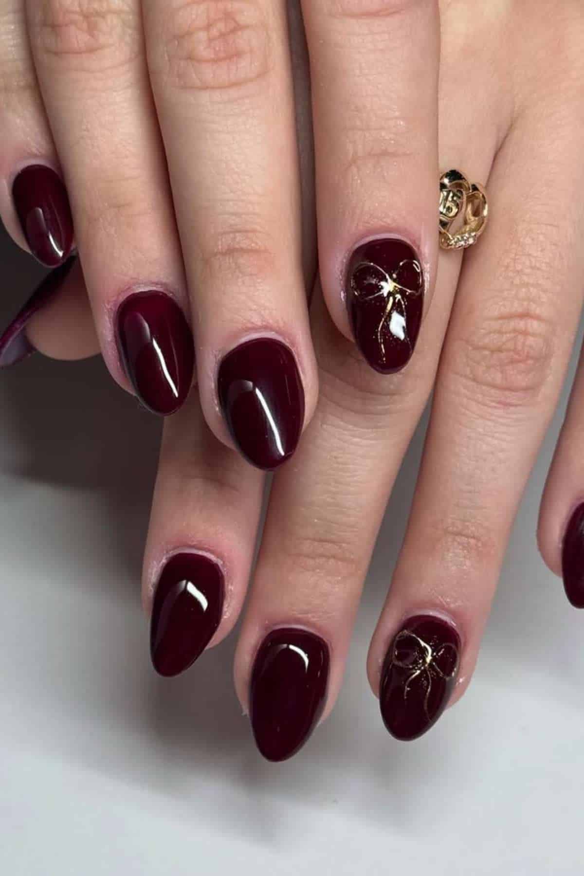 Hands with dark burgundy, almond-shaped nails; one nail on each hand features gold line art decoration. A gold ring is worn on one finger.
