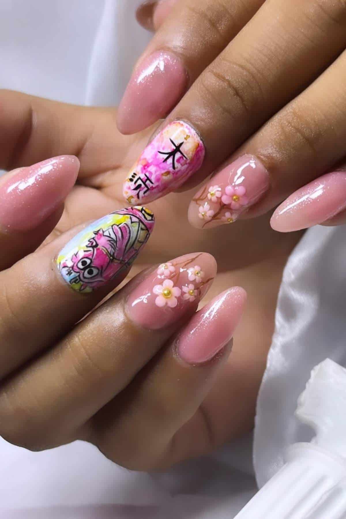 Hands with long, almond-shaped pink nails. Accent nails feature floral designs, Japanese characters, and an illustrated face.