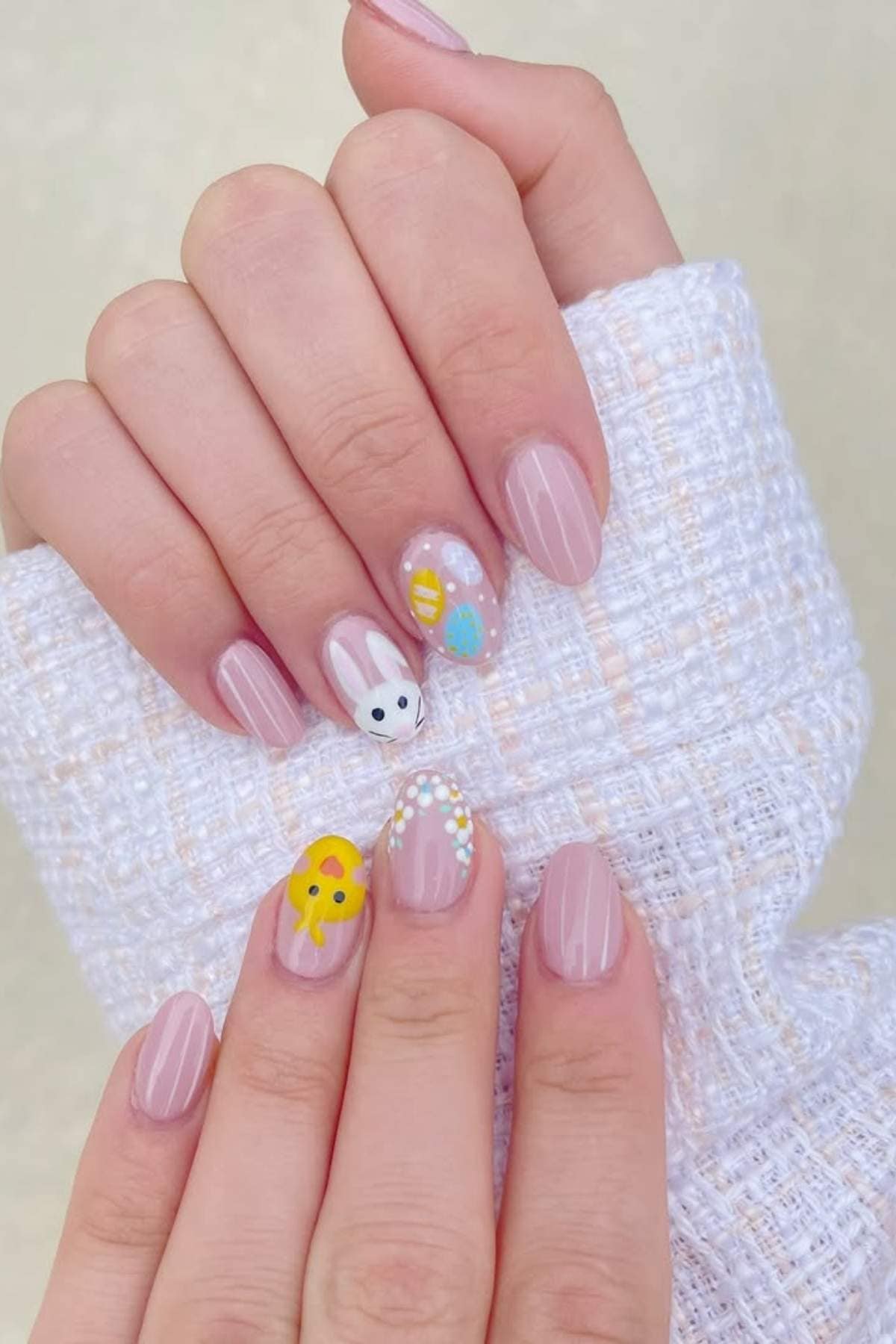 A hand with light pink manicured nails, featuring designs of a white bunny, colorful Easter eggs, and a yellow chick on two accent nails.
