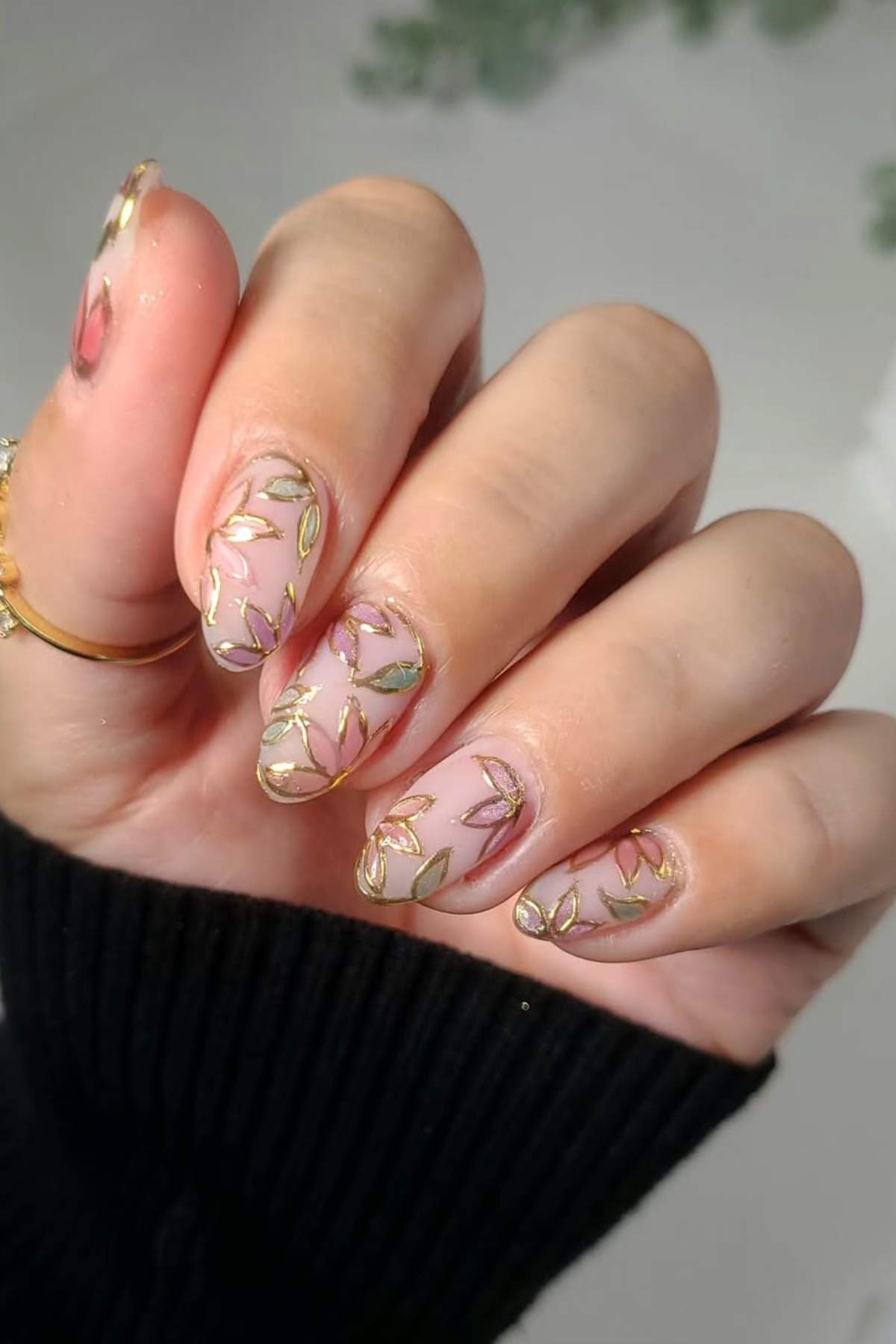 A hand with almond-shaped nails featuring a translucent base and gold-outlined leaf designs, shown against a blurred background.
