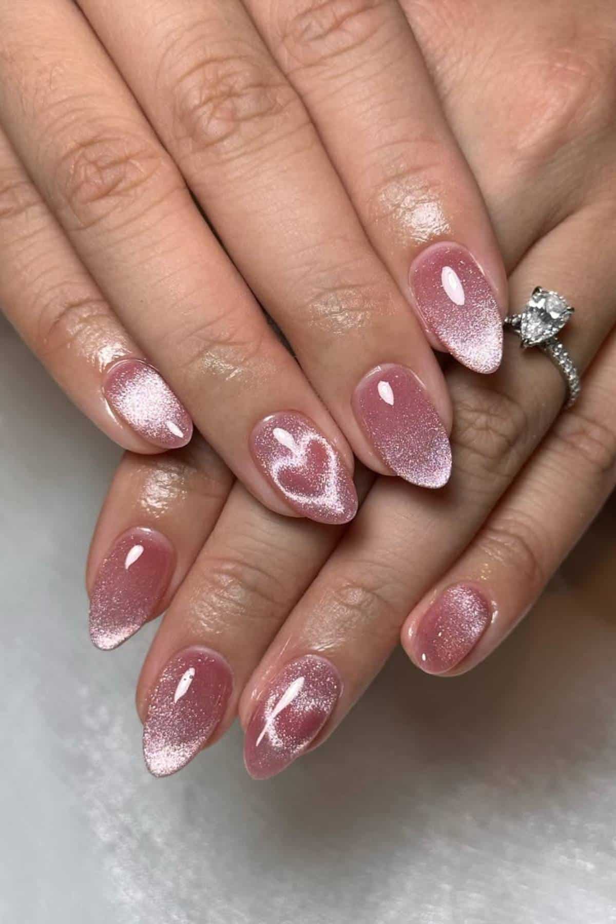Hands with almond-shaped nails painted in shimmery pink polish, featuring a subtle heart design on the ring fingers. A diamond ring is worn on the left hand.