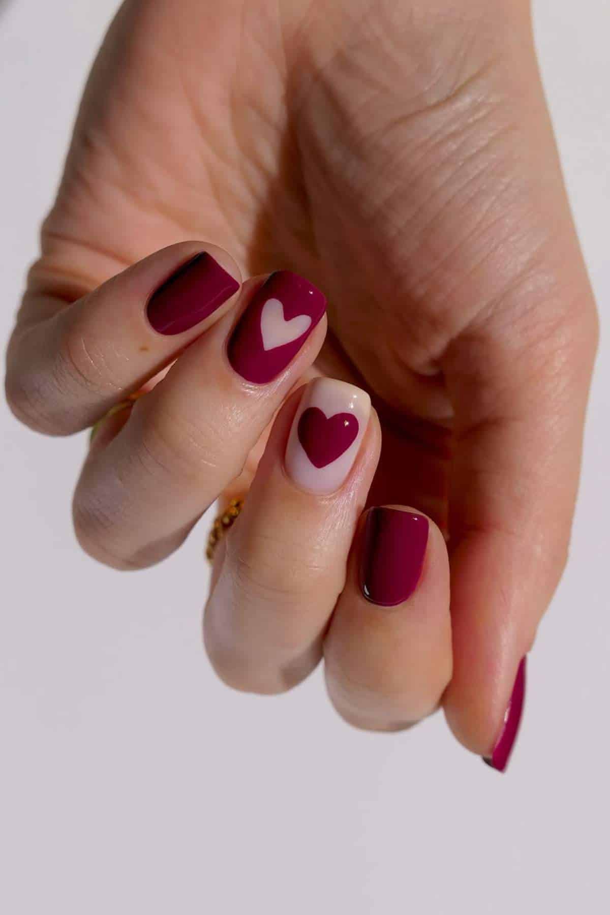 A hand with short, almond-shaped nails painted dark red; two nails feature white and red heart designs on a nude base.