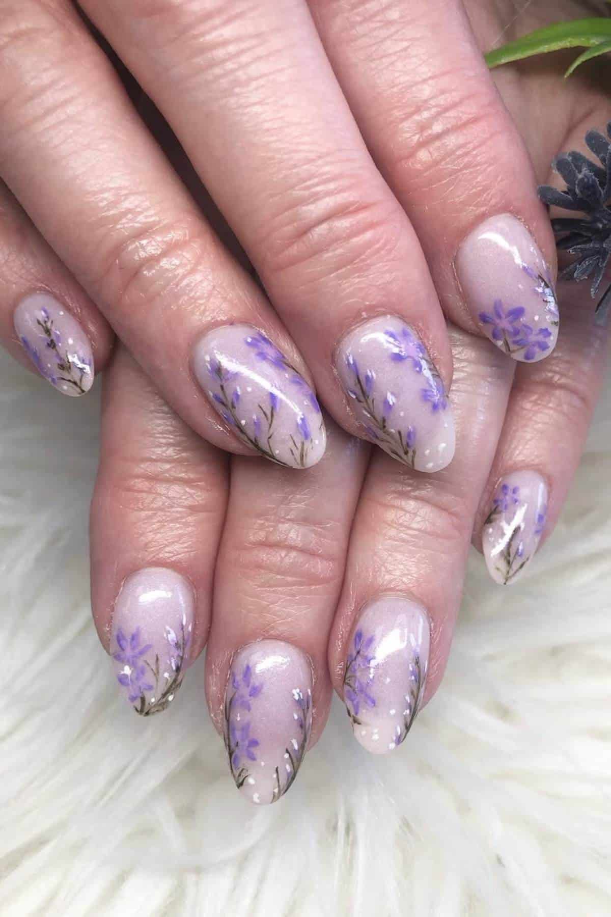 Hands with almond-shaped nails featuring a nude base color and purple floral designs with small branches and glitter accents.