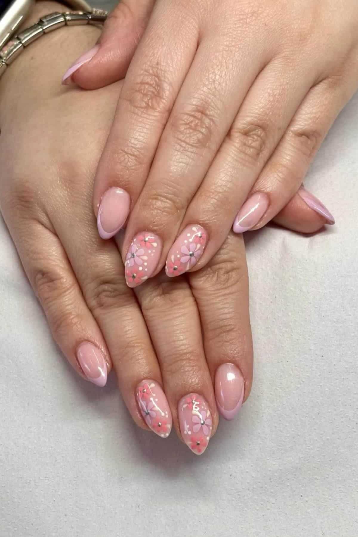 Hands with almond-shaped nails painted in light pink polish, some featuring delicate floral designs in white and purple.