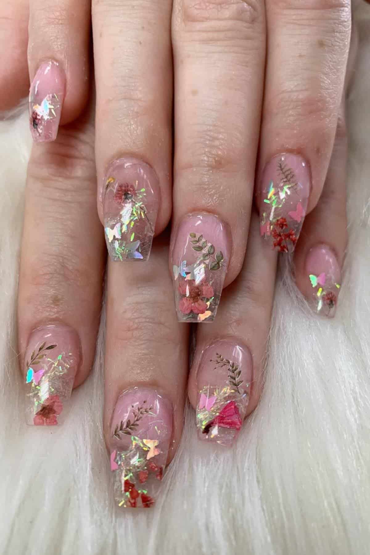 Close-up of hands with long, square-shaped nails featuring clear polish, dried flowers, gold accents, and iridescent flakes as nail art, resting on a white furry surface.