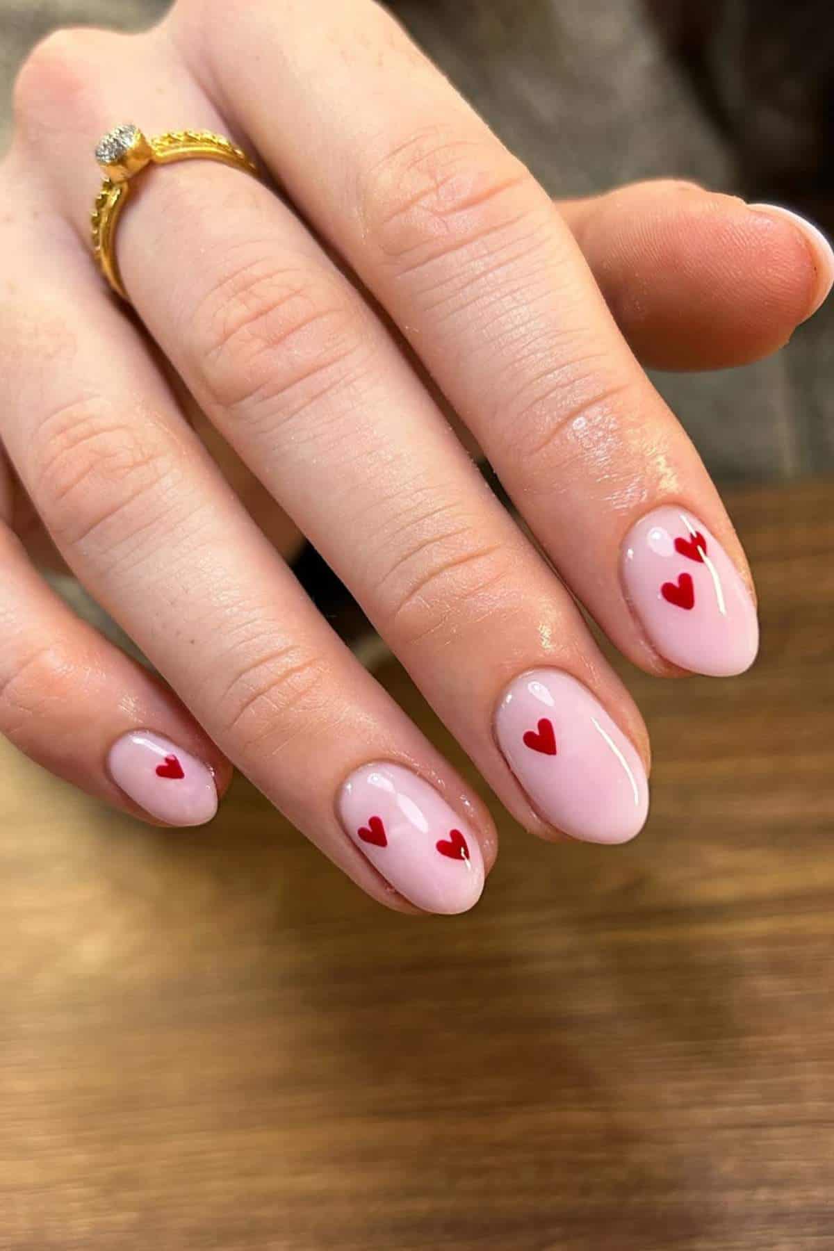 A hand with light pink nail polish and small red heart designs on each nail is shown resting on a wooden surface. The person is wearing a gold ring.