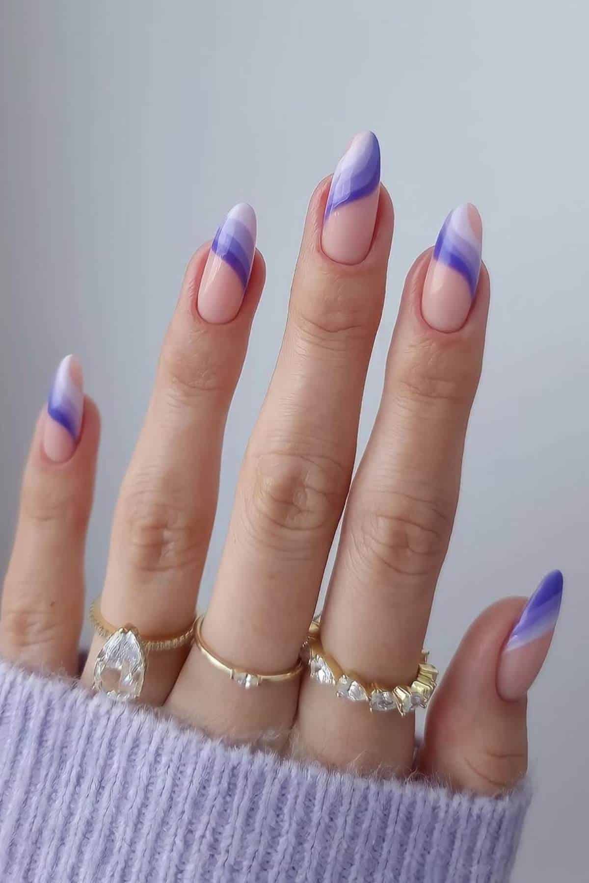 A hand with almond-shaped nails features purple and lavender diagonal swirl designs. The person wears gold rings and a lilac knitted sleeve is partially visible.