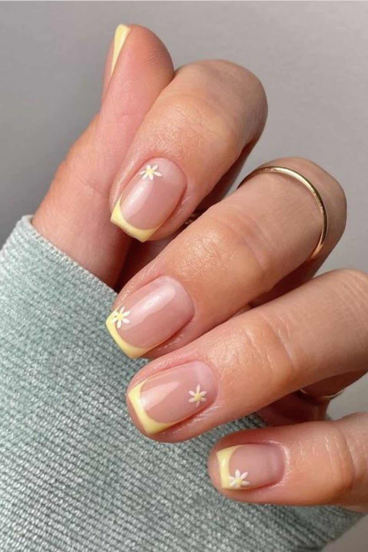 A hand with short, square nails featuring yellow French tips and small white daisy designs on a light pink base.