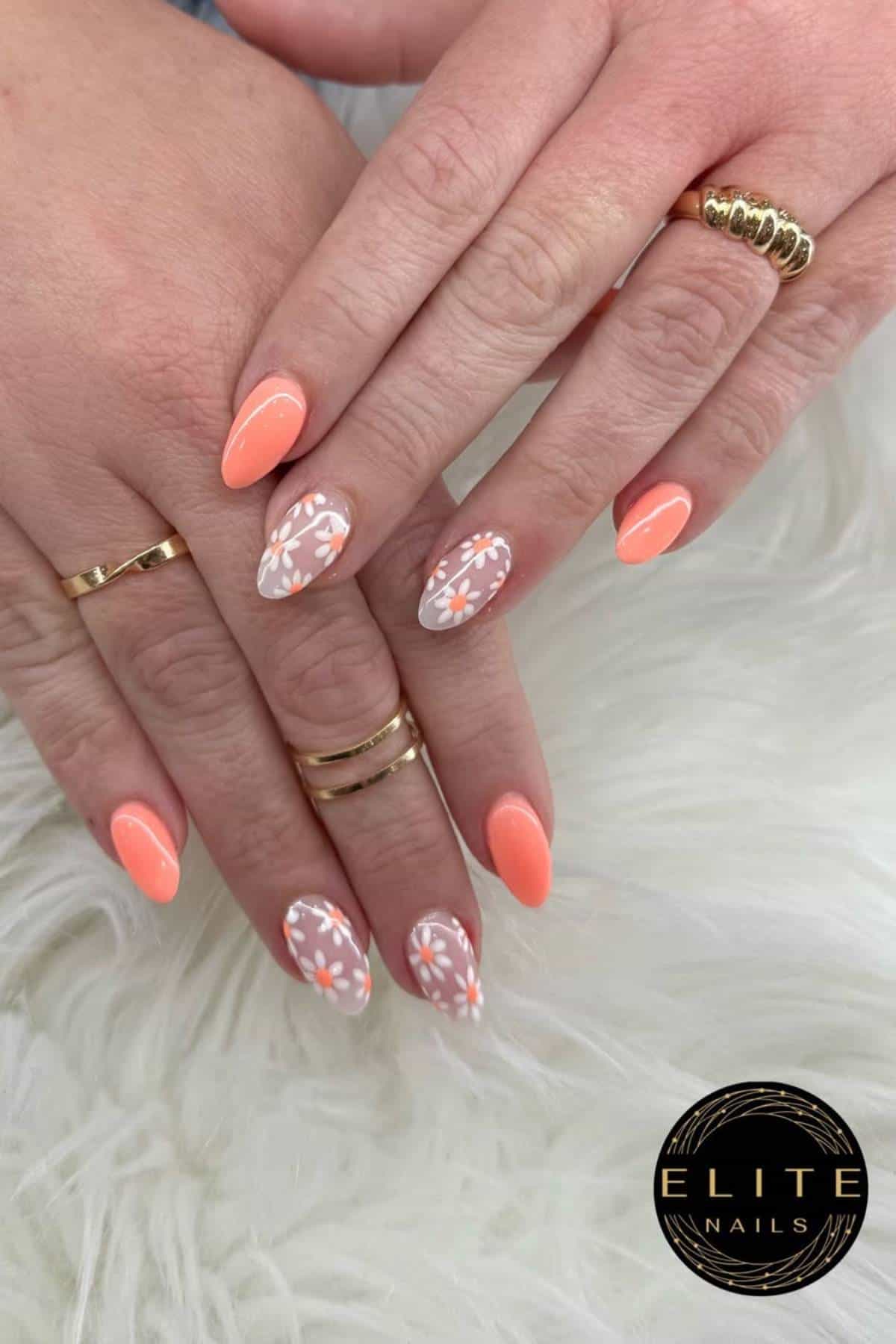 Hands with almond-shaped nails, painted orange with two accent nails featuring white and orange floral designs, placed on a white textured surface. "ELITE NAILS" logo in the corner.