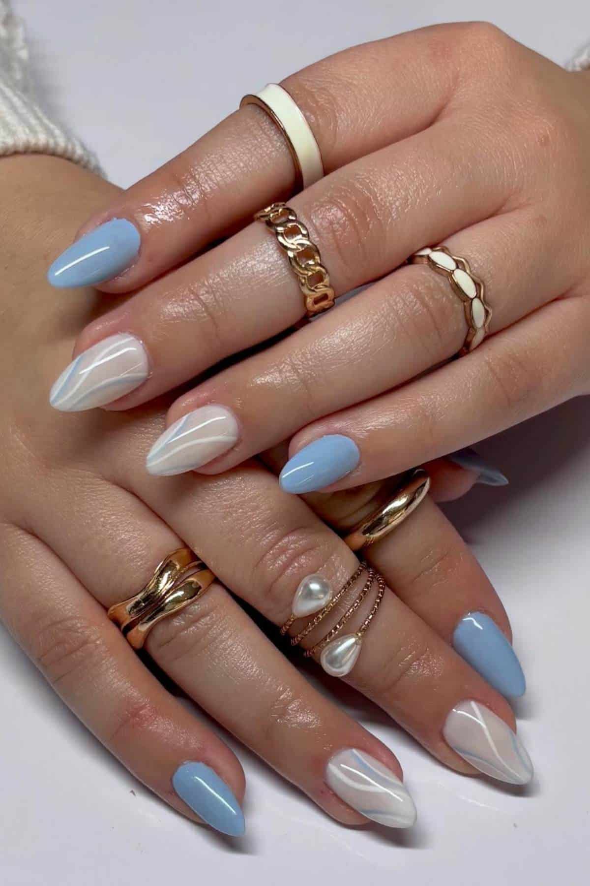 Hands with almond-shaped nails painted in pastel blue and white swirl designs, wearing multiple gold and white rings on each finger.