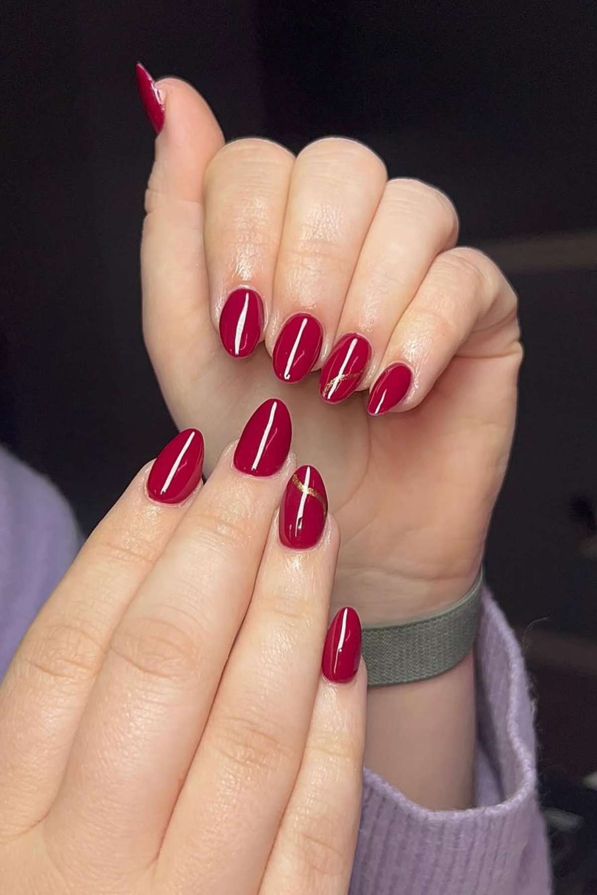Hands with oval-shaped nails painted glossy dark red; one nail on each hand features a thin gold diagonal line as an accent.