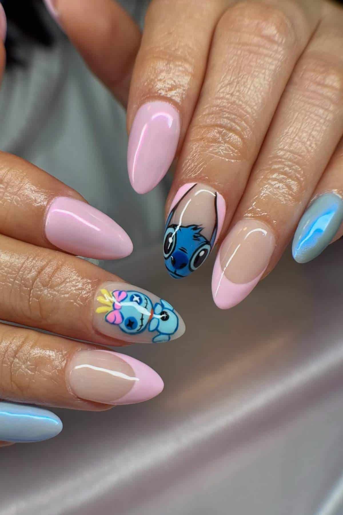 Hands with long almond-shaped nails painted in pastel pink, blue, and nude colors; two nails feature cartoon character designs with detailed faces.