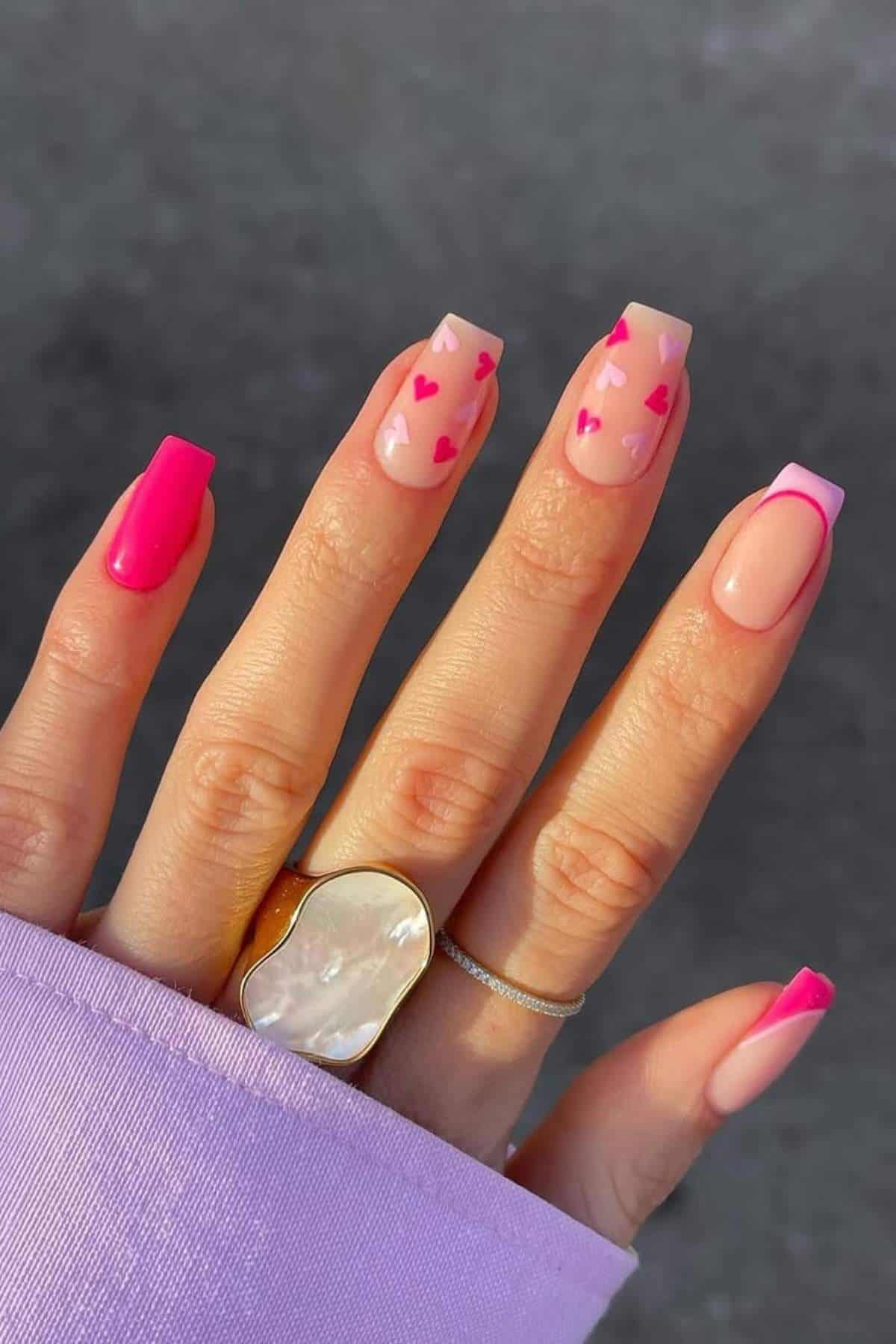 A hand with long square-shaped nails; some are painted pink, some have pink heart designs, and one has a pink French tip. The person wears a gold ring and a lilac sleeve.