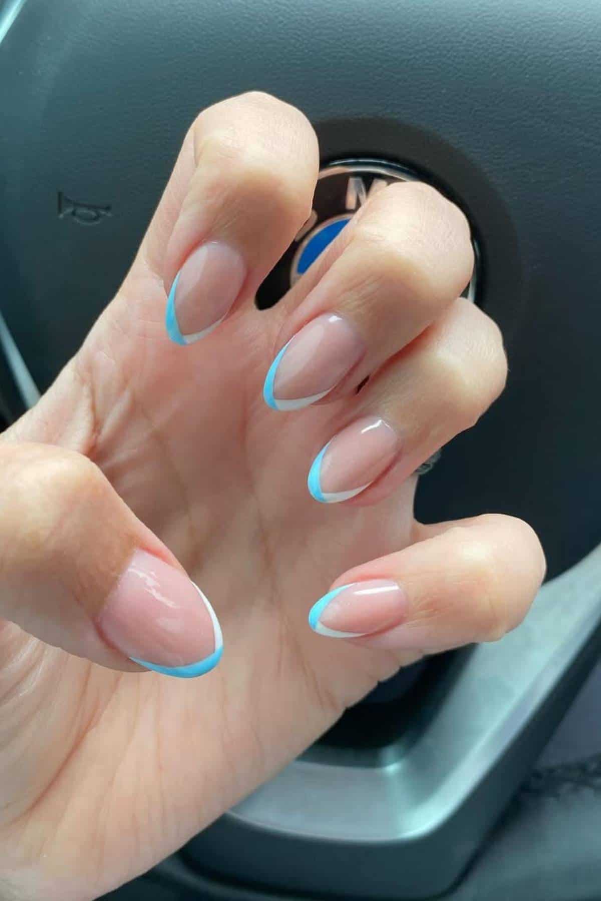 A hand with neatly manicured nails featuring a light blue French tip design, shown in front of a car steering wheel.