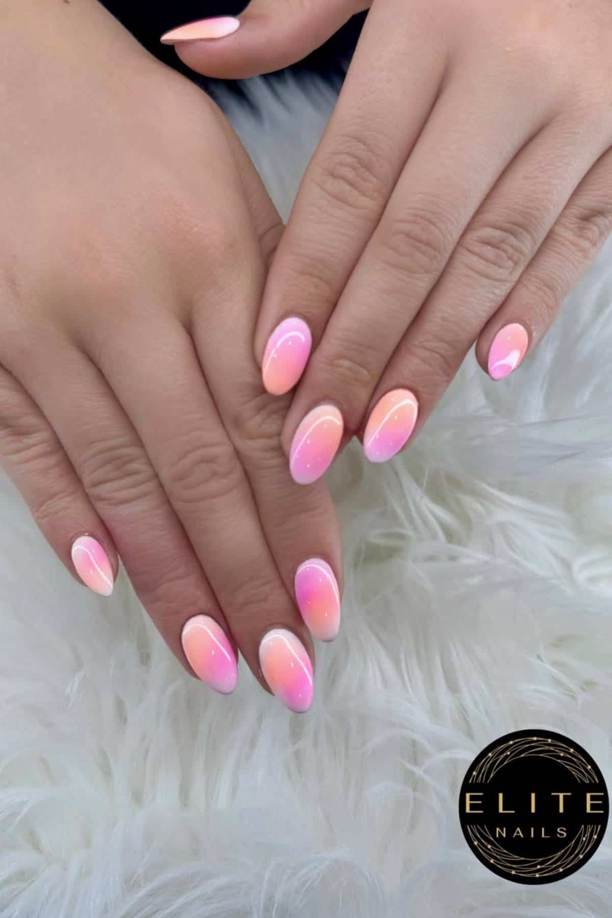 Hands with almond-shaped nails painted in a pink and peach ombré design with a glossy finish, shown on a white furry surface. "Elite Nails" logo is visible in the bottom right corner.