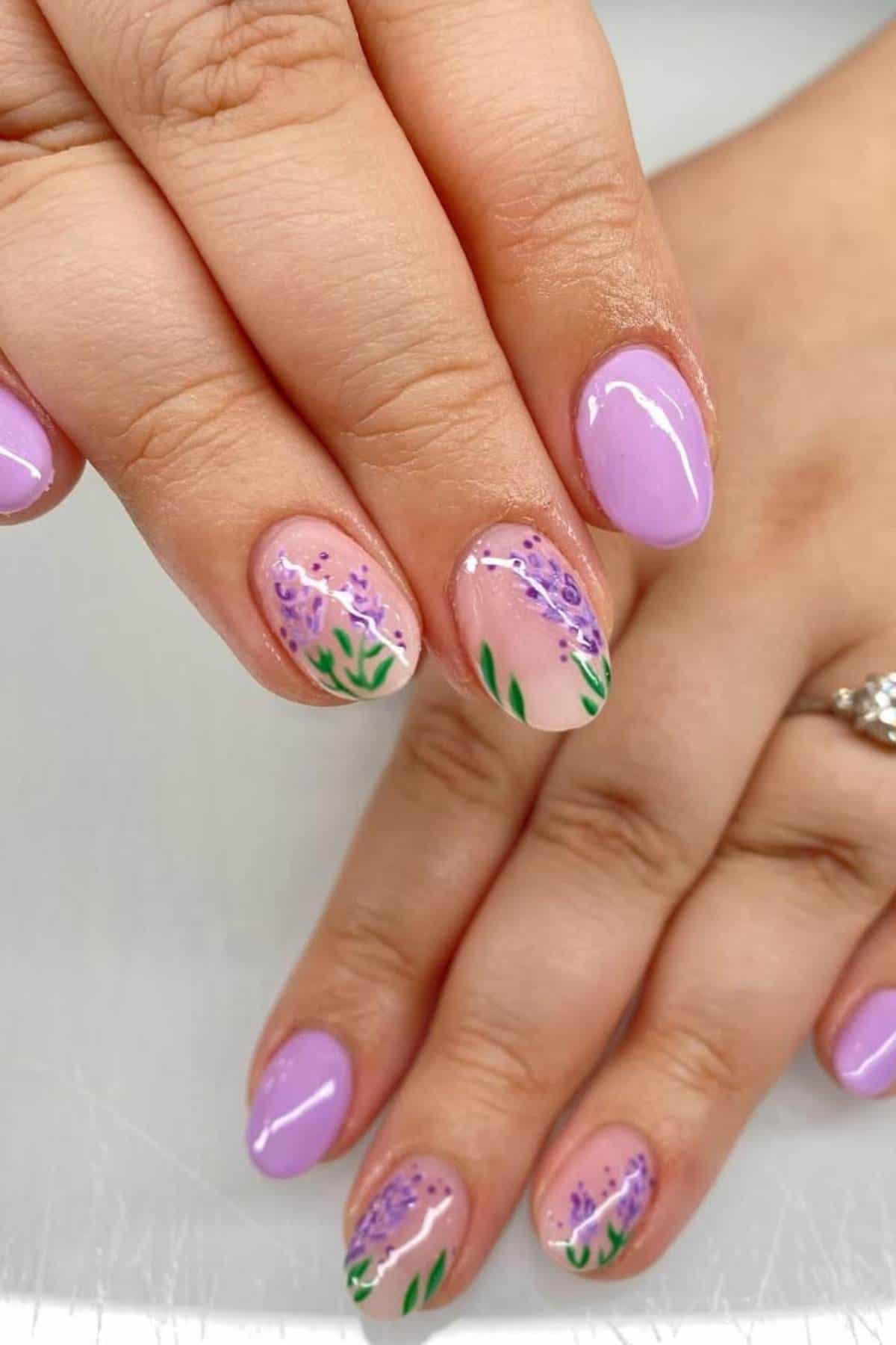 Hands with oval-shaped nails painted in light purple polish; two nails feature hand-painted lavender flower designs with green stems on a nude base.