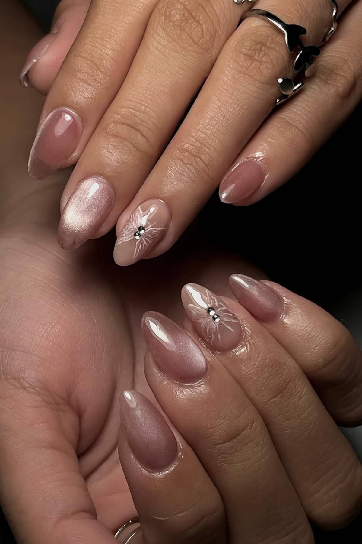 Hands with almond-shaped nails featuring a glossy nude-pink polish, accent nails with white floral designs, and small rhinestone details. Rings are worn on some fingers.