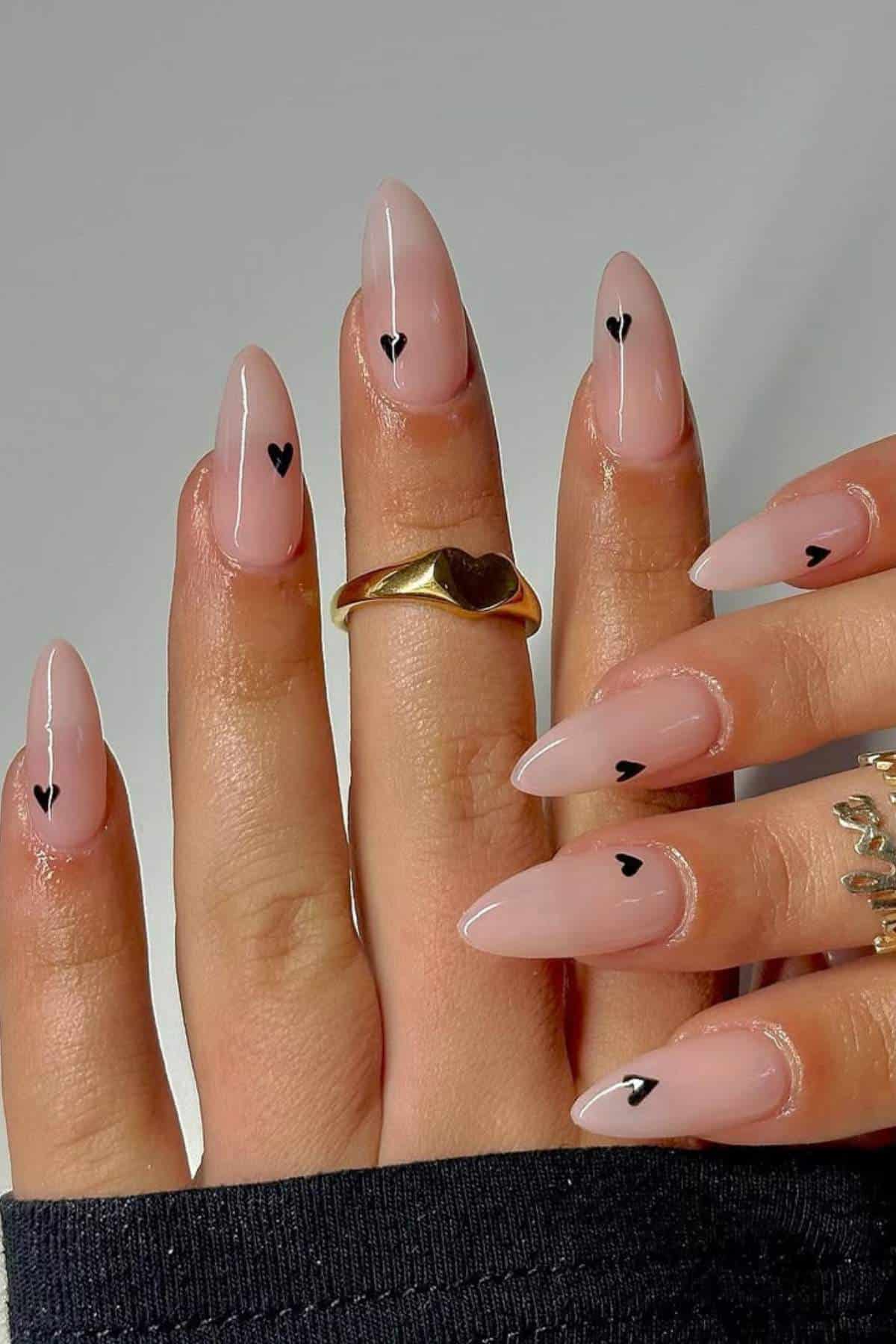 A hand with long, almond-shaped nails featuring a glossy nude base and small black heart designs near the cuticle on each nail. The person wears gold rings.