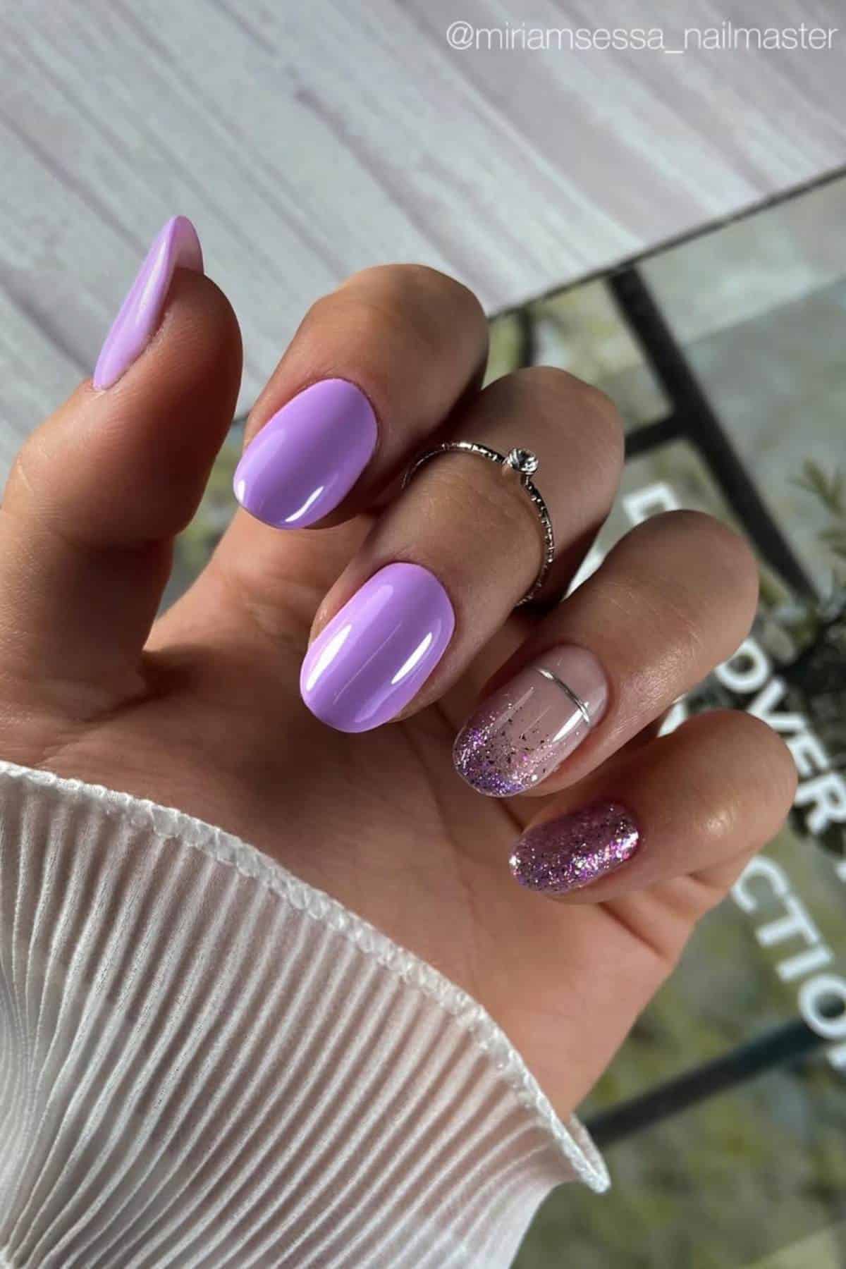 A hand with almond-shaped nails painted in light purple; one nail is clear with glitter tips, and another nail is fully covered in purple glitter. The hand wears a silver ring.